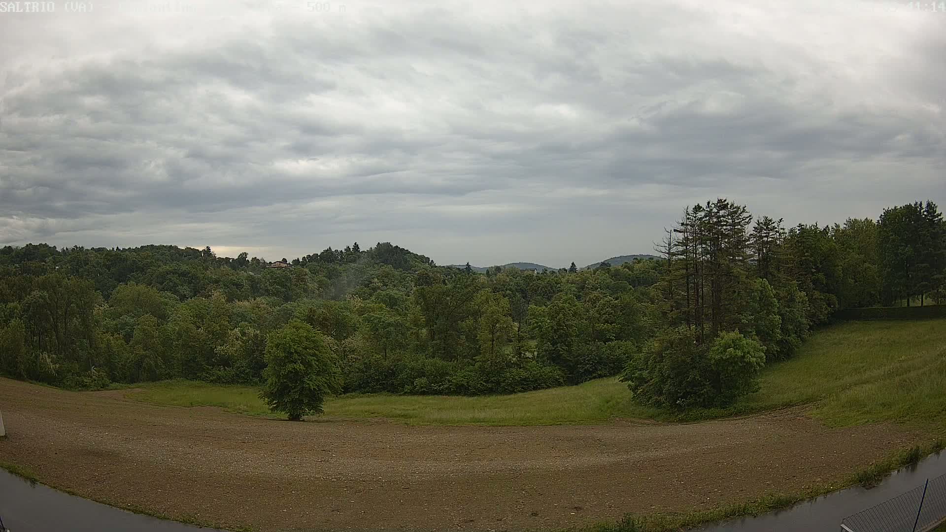 A mostly cloudy sky hangs over a verdant landscape of trees and fields, with a section of bare, brown earth in the foreground.