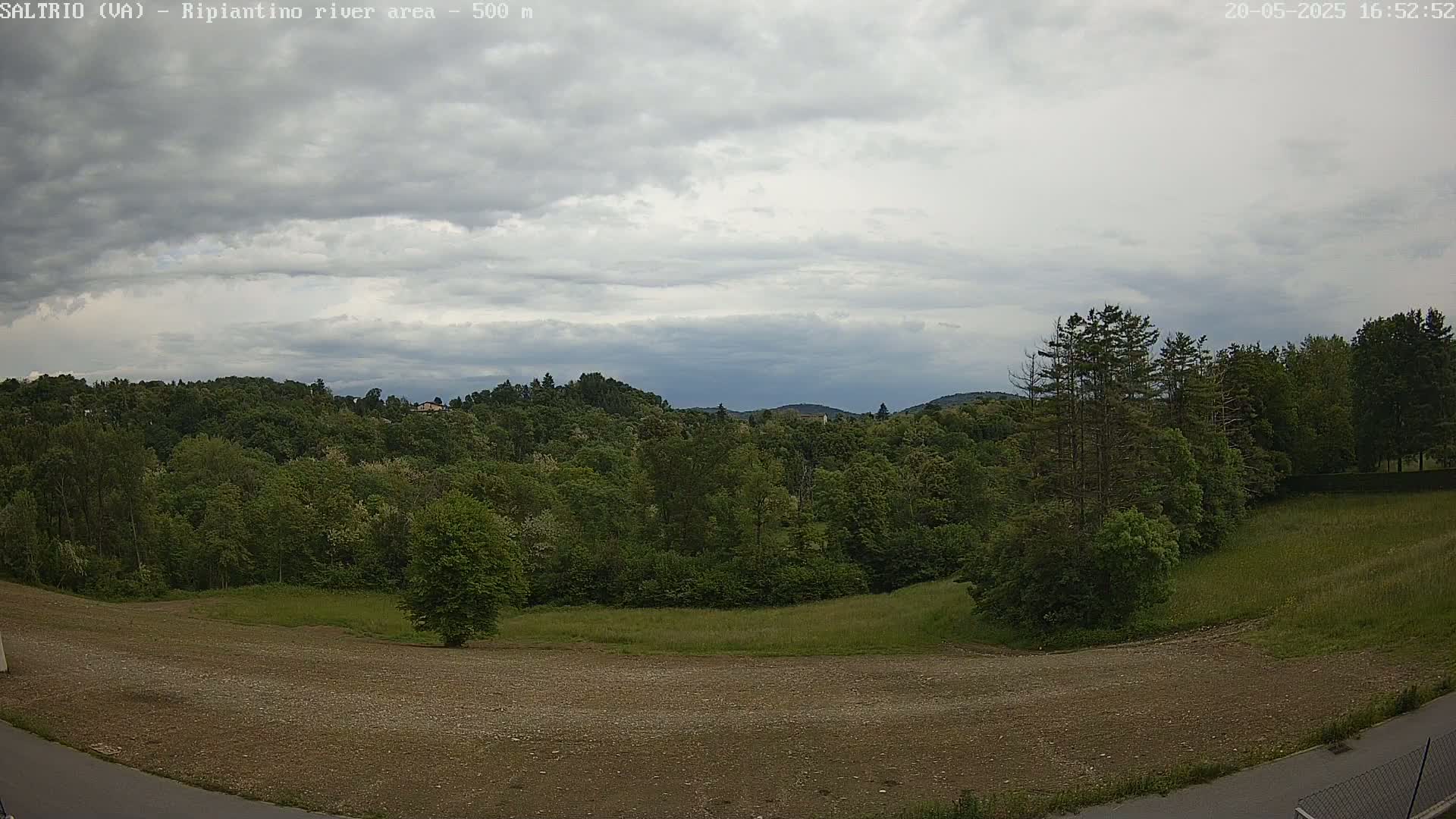 A mostly cloudy sky overlooks a grassy field and a treeline of lush green deciduous trees on a hill, with a barren, dirt area in the foreground.