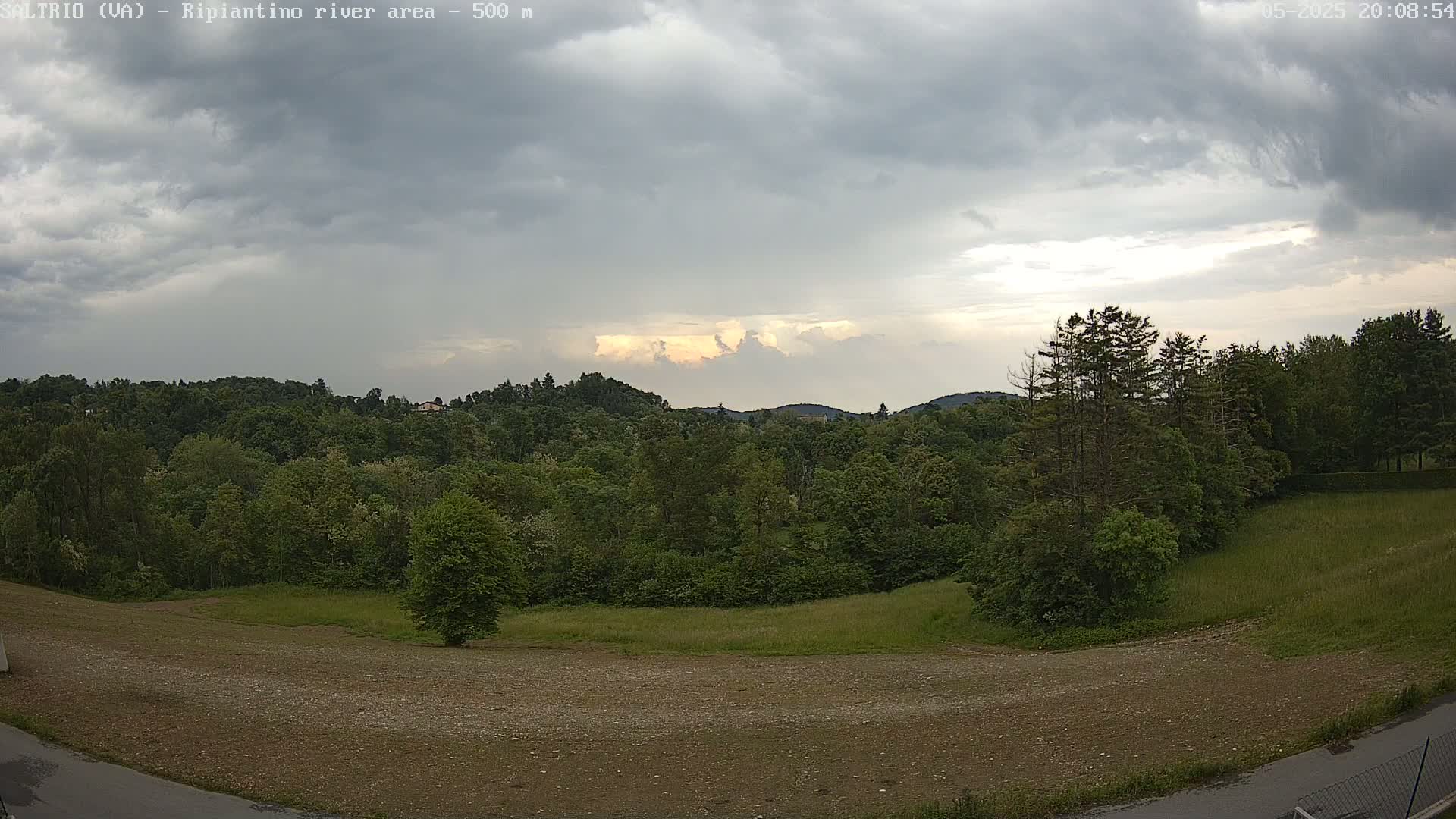 A mostly cloudy sky with a break of lighter clouds above a line of green trees and a brown, grassy field.
