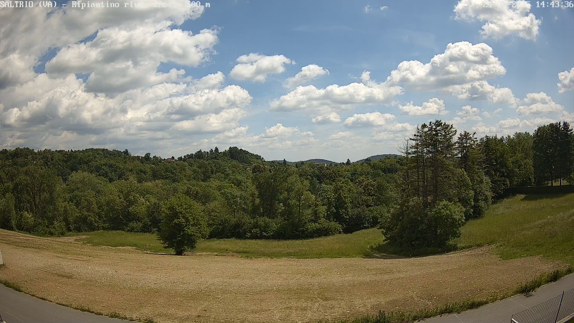 A partly sunny day shows a view of a plowed field, green grassy areas, and a lush green forest with hills in the background.