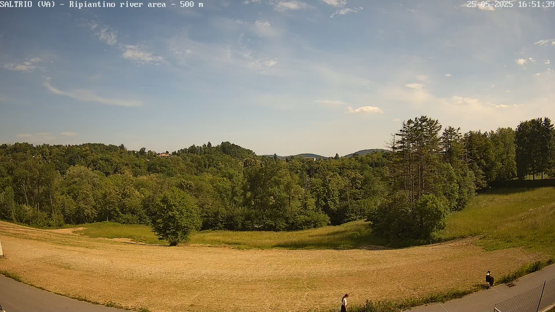 A mostly sunny day reveals a hilly landscape of lush green trees and a tan, sparsely vegetated field, with two people walking near a paved road in the bottom right.
