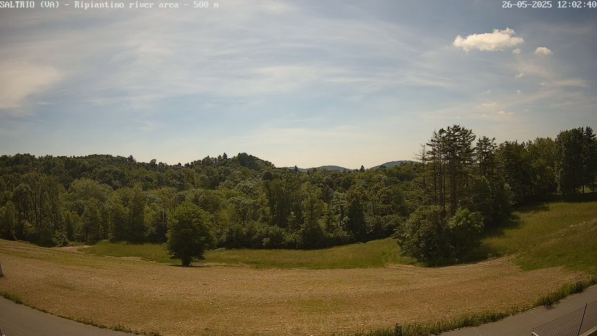 A mostly sunny day reveals a view of a verdant, hilly landscape with a mix of trees and a brown, open field in the foreground.