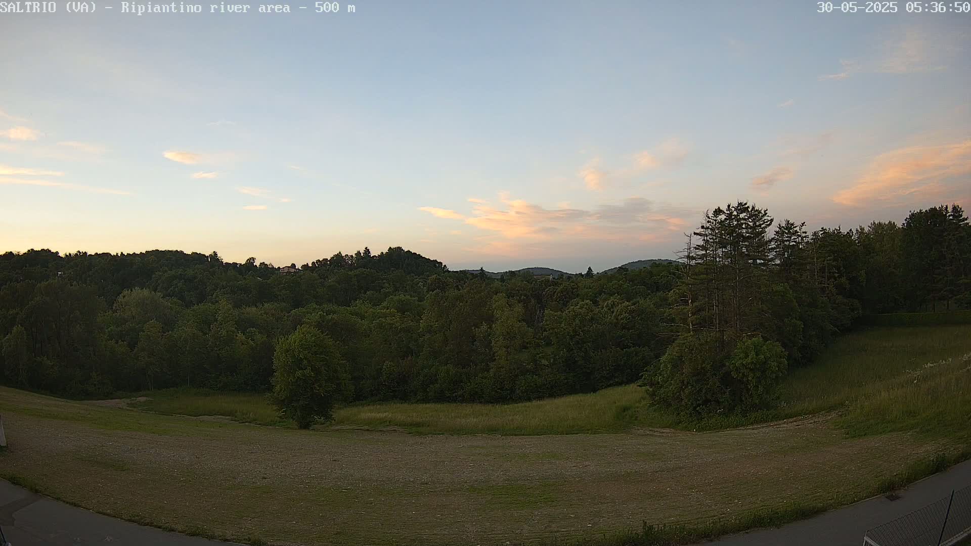 A high-angle view of a verdant landscape at sunset, featuring a hilly terrain covered in lush green trees and an open field, under a light pink and blue sky with wispy clouds.