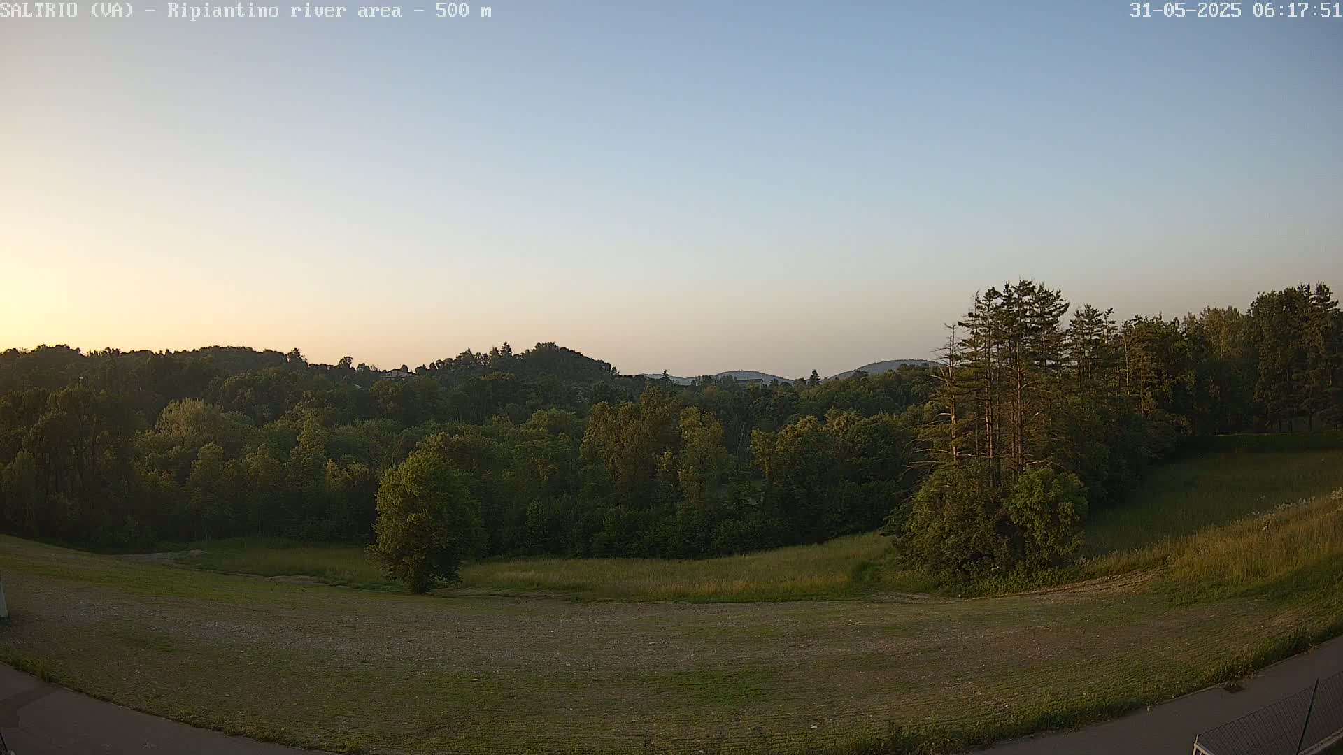A high-angle, long shot shows a tranquil, verdant landscape under a clear, pale-blue early morning sky.