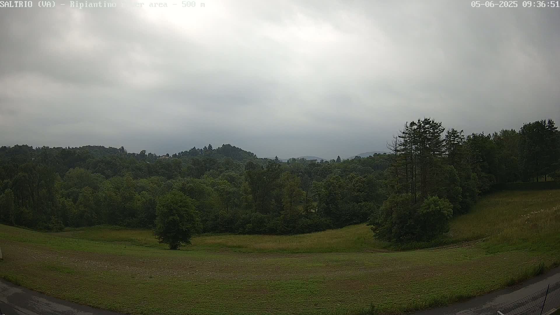 A mostly overcast sky hangs over a verdant landscape of rolling hills and fields dotted with lush green trees.