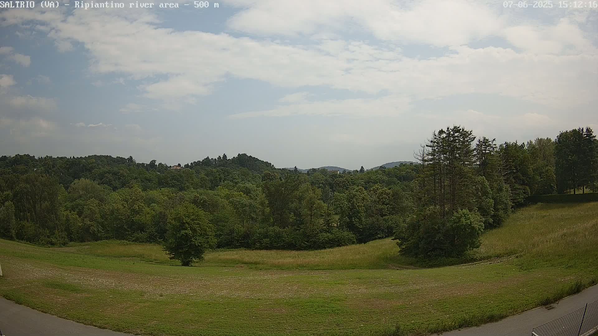 A partly cloudy sky overlooks a verdant landscape of rolling hills covered in lush green trees and grassy fields.
