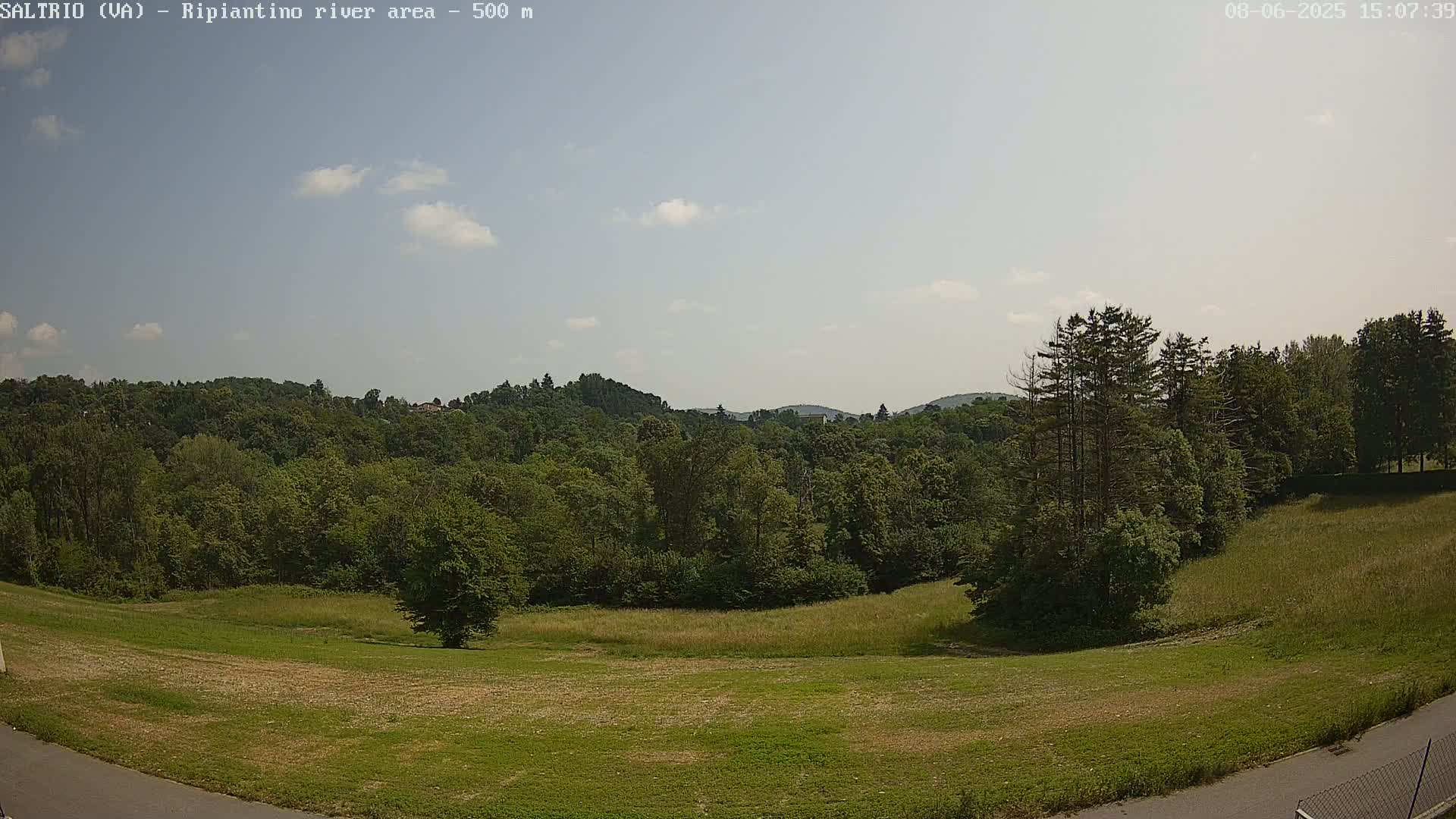 A mostly sunny day reveals a verdant landscape of grassy fields and lush green forests, rolling hills in the background.