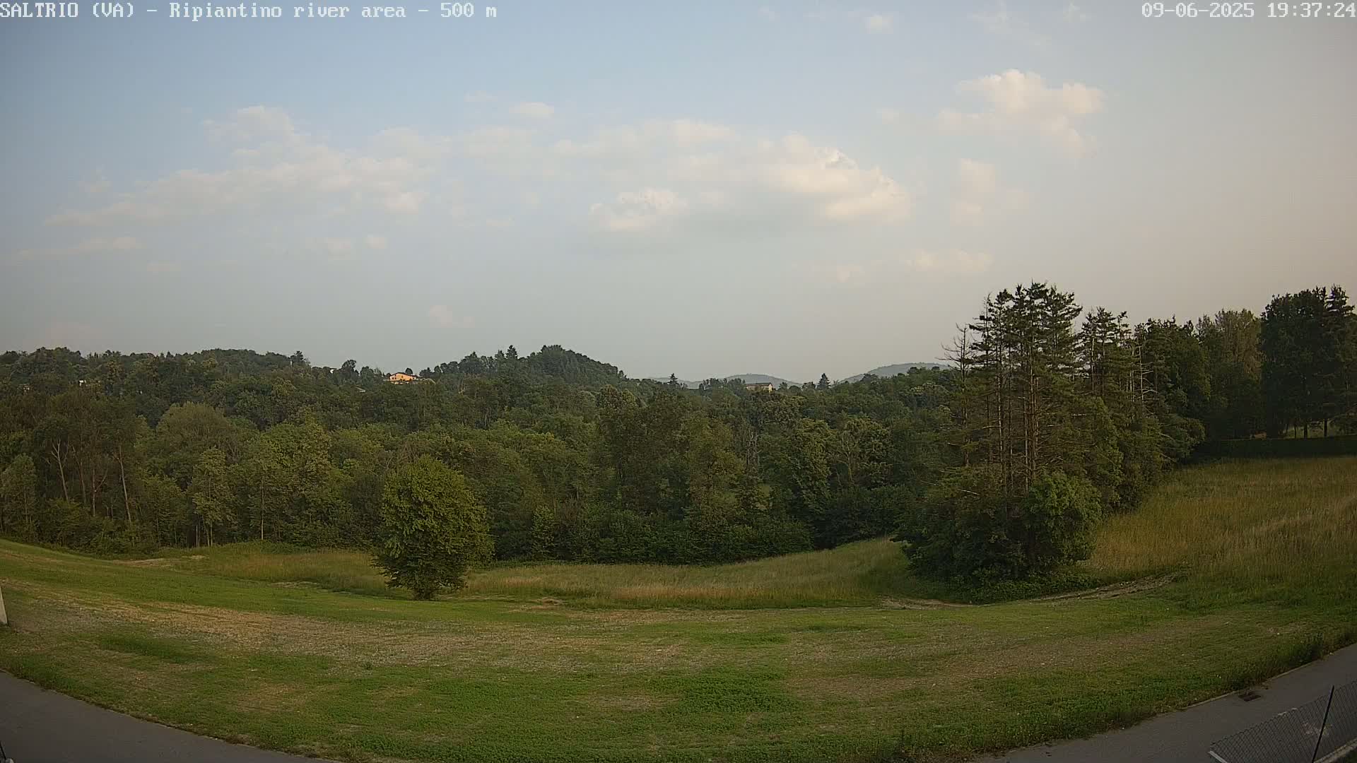 A partly cloudy sky overlooks a wide expanse of green fields and forest, with a few tall trees standing prominently near the right.