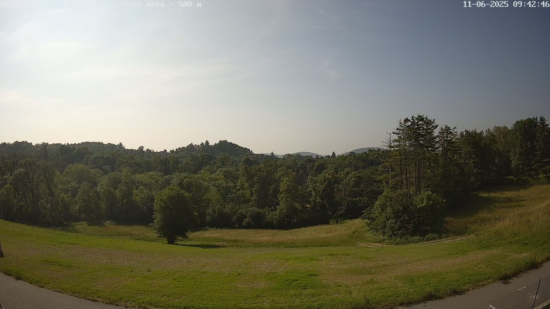 A mostly clear sky overlooks a grassy field and a dense forest covering rolling hills in the distance.