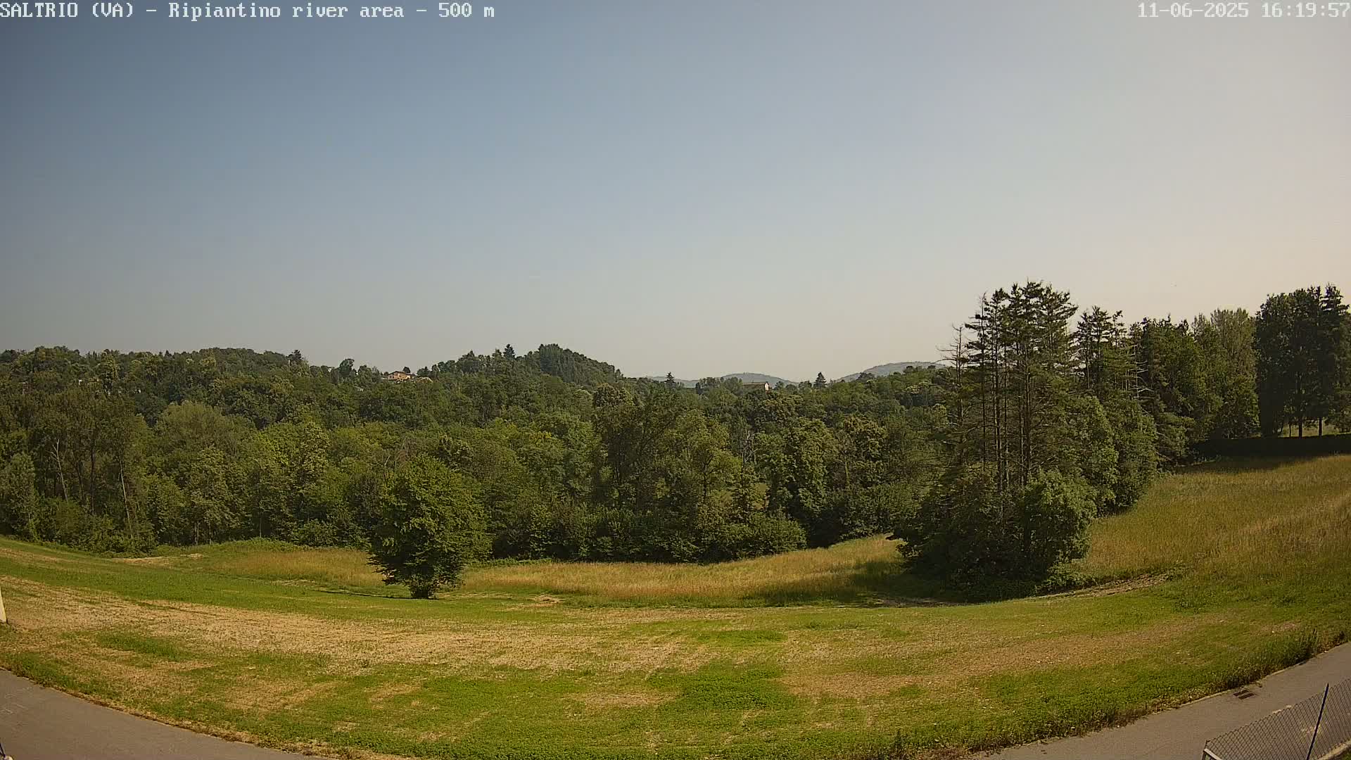 A mostly sunny day reveals a grassy field sloping down to a line of lush green trees and distant hills under a pale blue sky.
