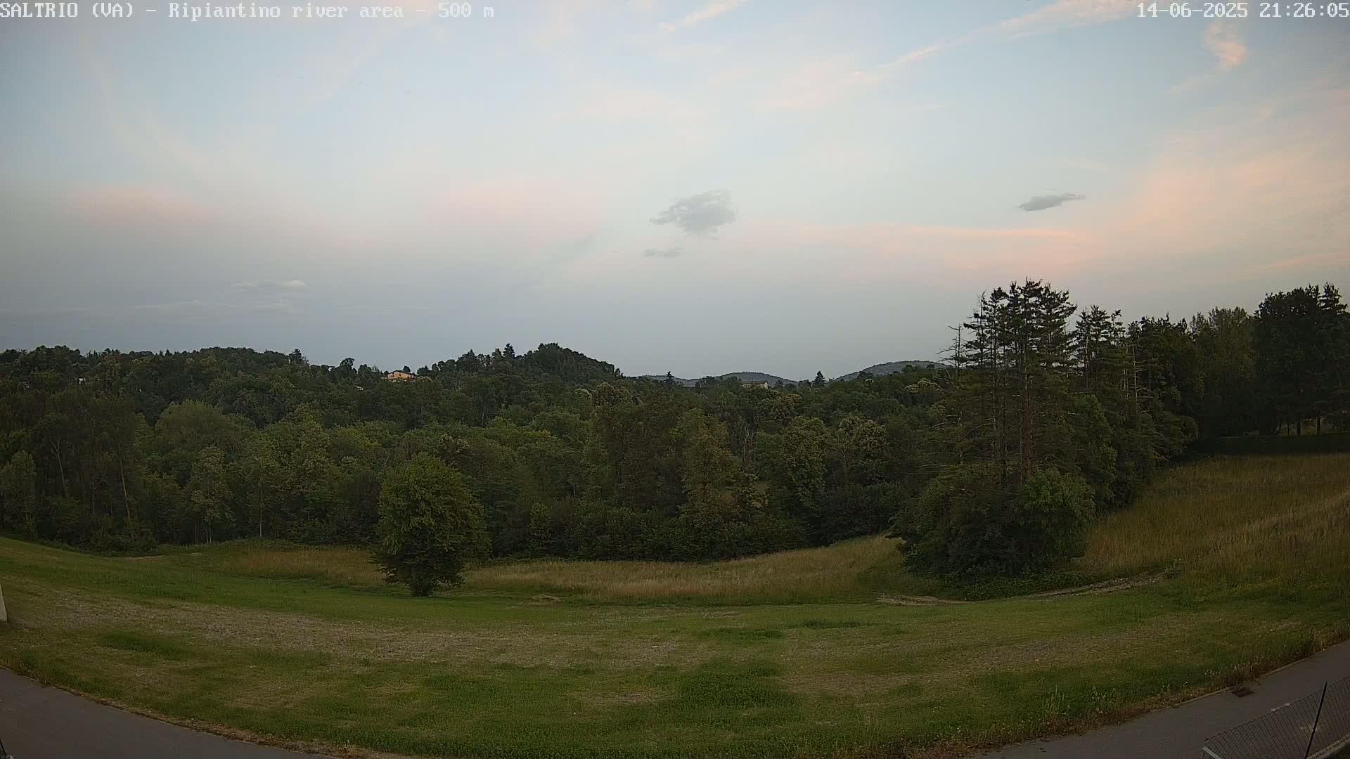 A mostly cloudy sky overlooks a verdant landscape of rolling hills covered in lush green trees and fields.