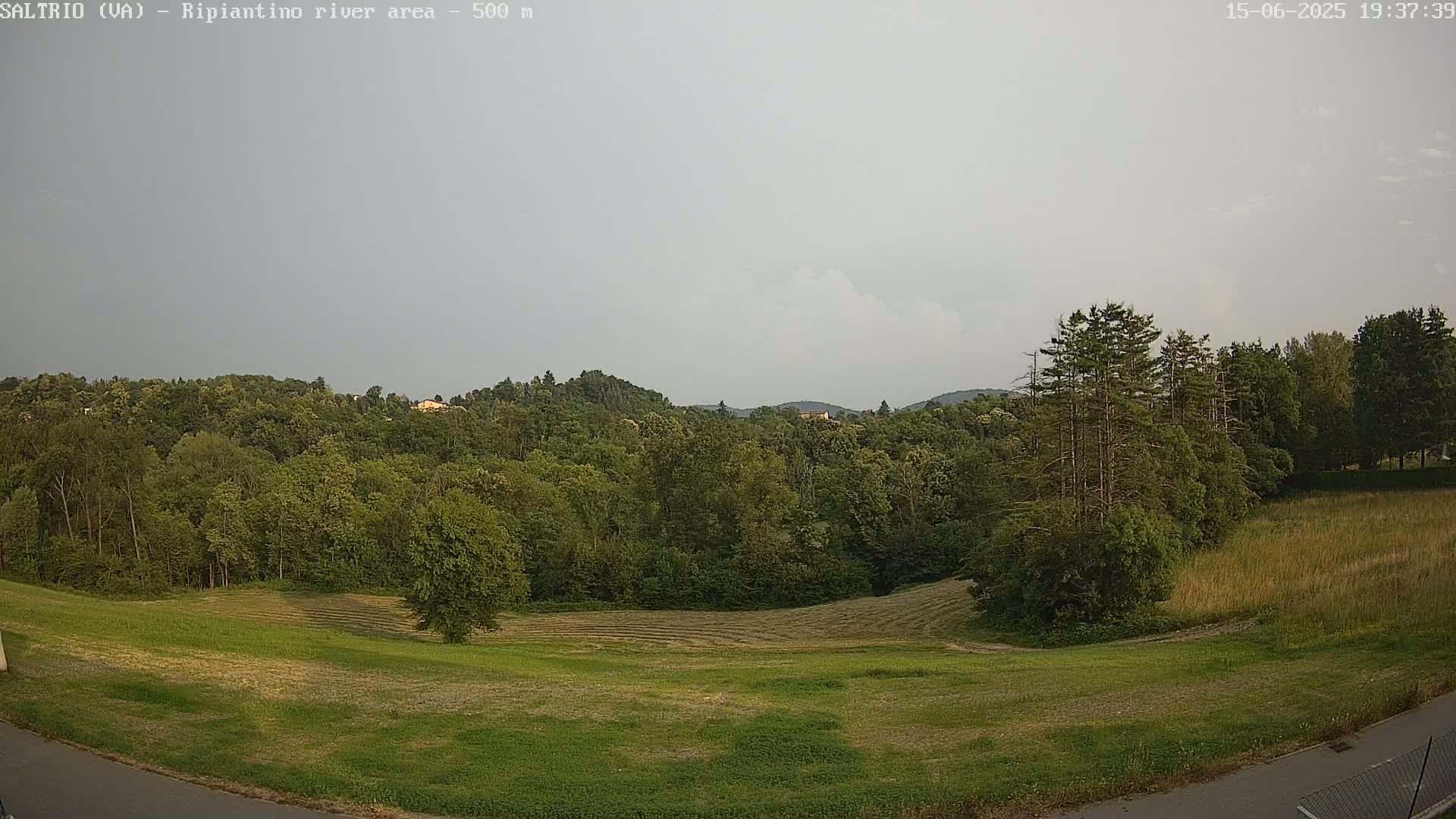 A mostly cloudy sky overlooks a lush green landscape of trees and mown fields, with taller trees near the right edge.