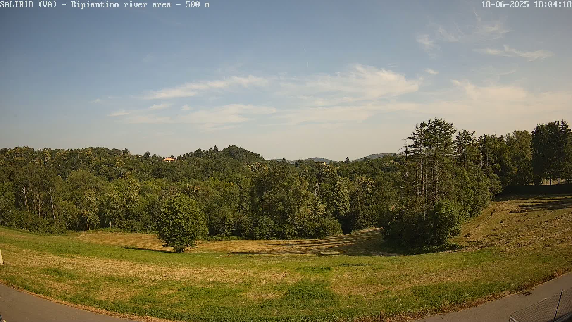 A mostly sunny, clear sky overlooks a verdant landscape of fields and a dense forest, with hills visible in the distance.