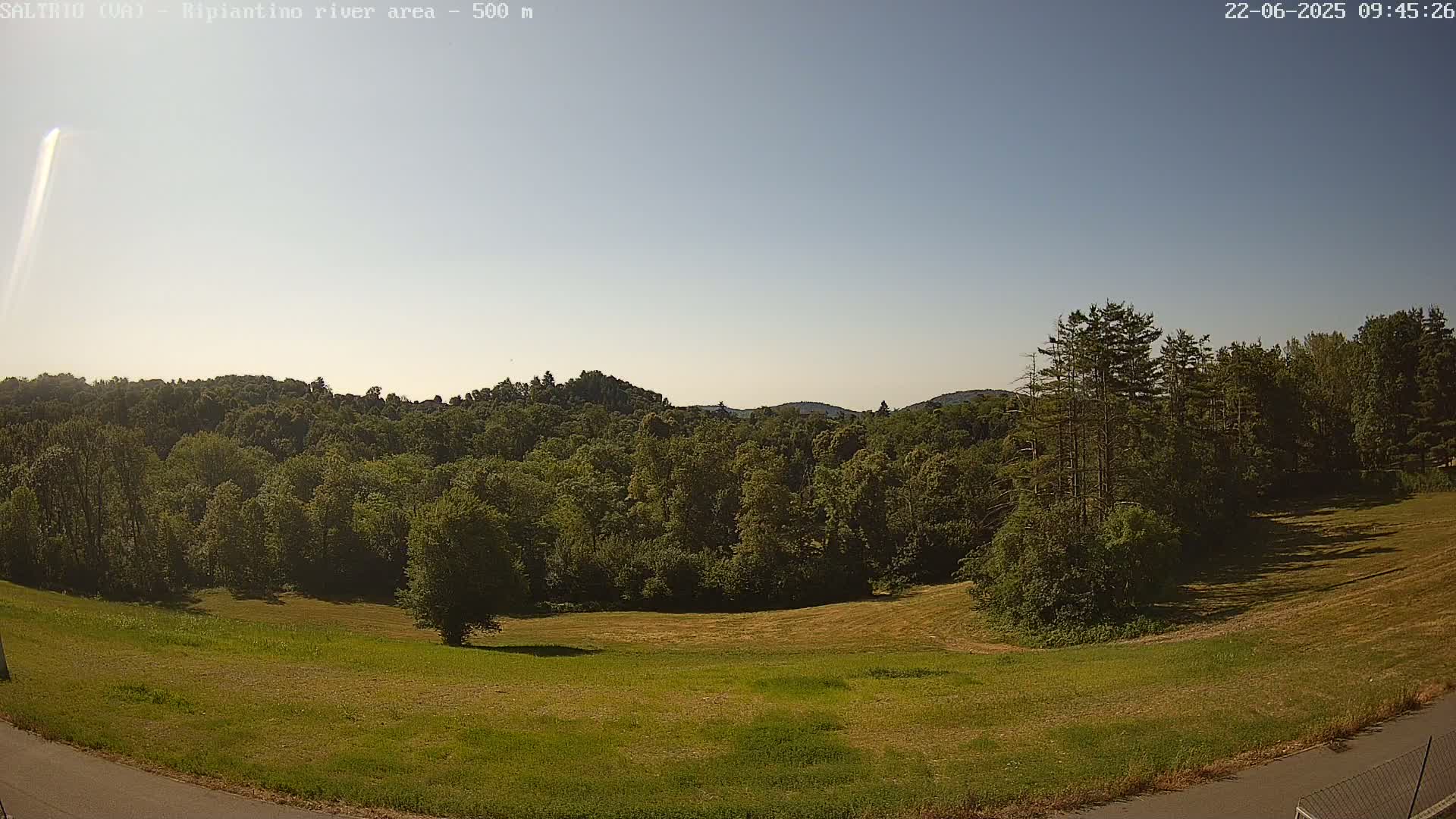 A mostly sunny day reveals a grassy field sloping down to a line of lush green trees and distant hills under a clear, light blue sky.
