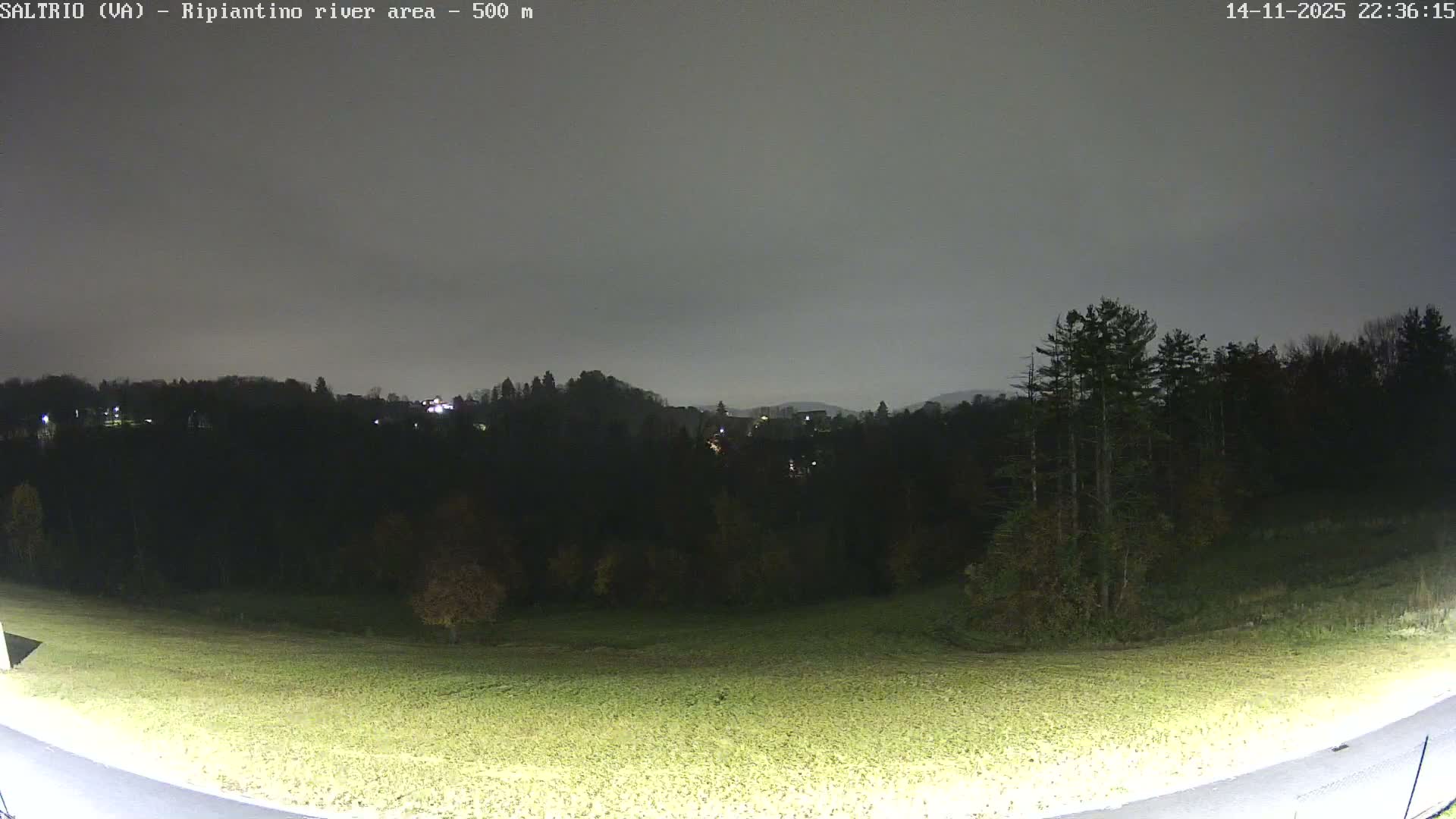 An outdoor night scene under a cloudy sky shows an illuminated grassy field in the foreground, with dark forested hills and scattered distant lights of buildings in the background.