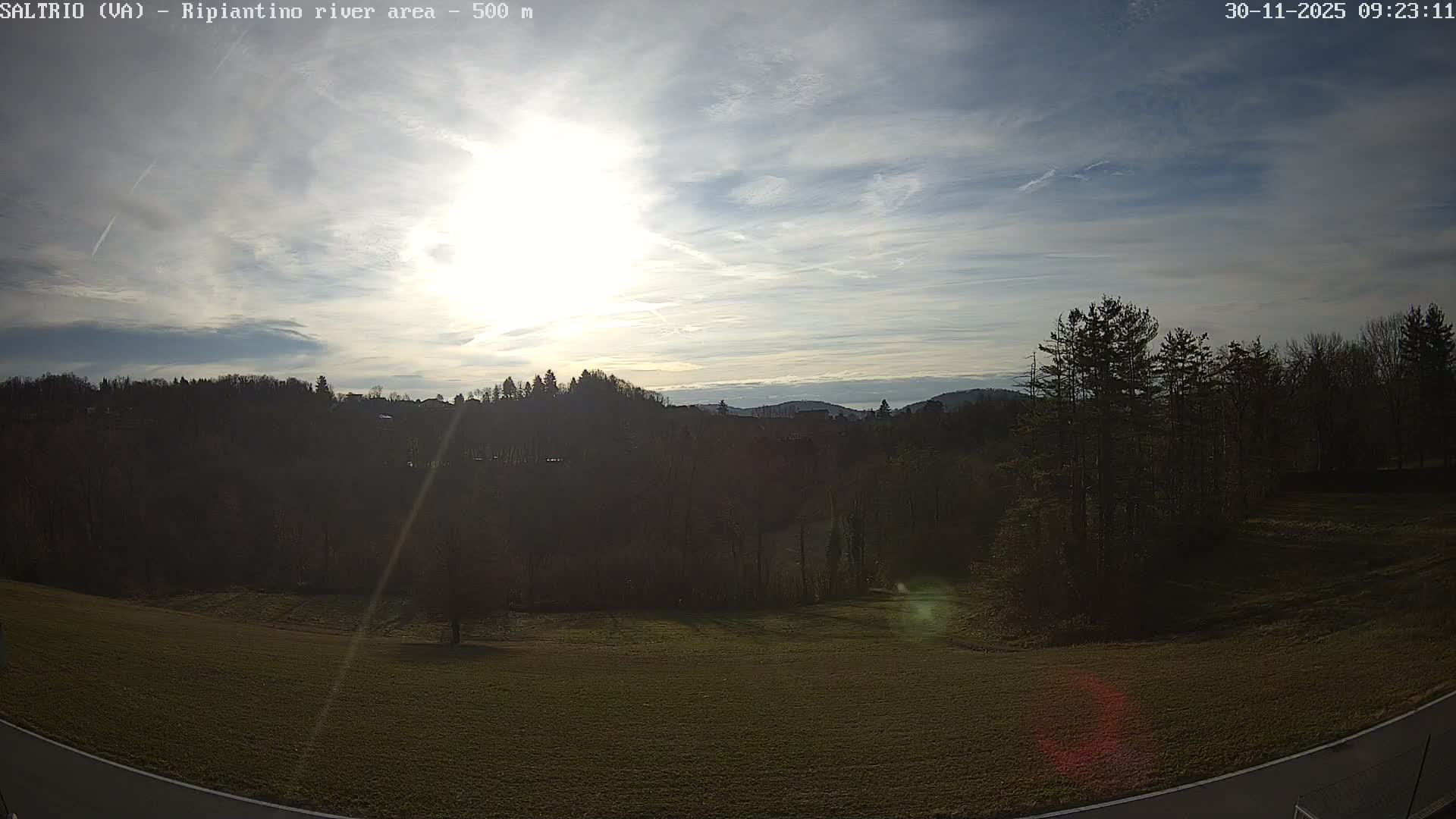 On a bright, partly cloudy morning, a wide outdoor landscape features a sun-drenched green field in the foreground, leading up to a dark forested hillside with distant hazy mountains under a sky scattered with wispy clouds and a bright sun.