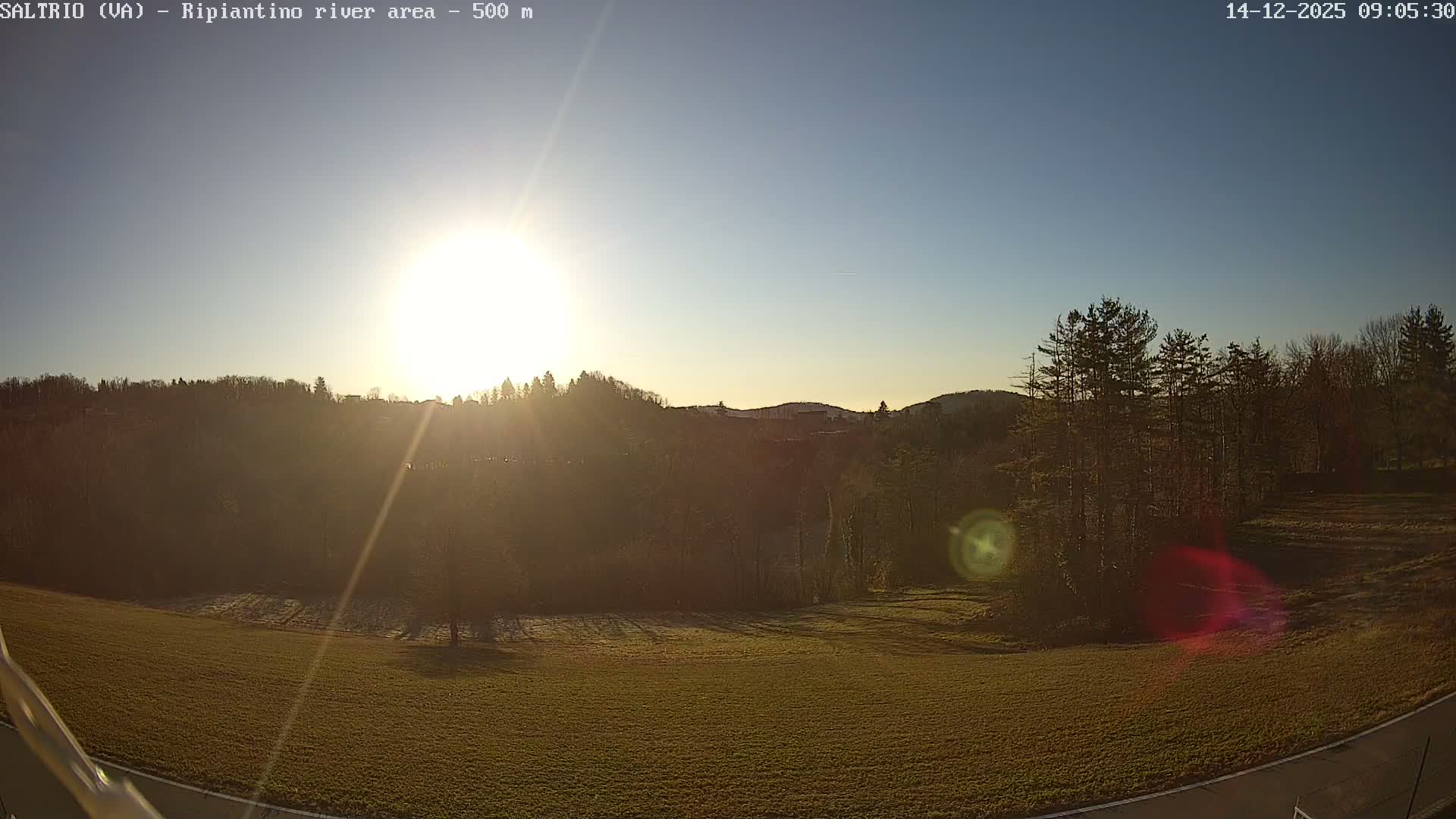 Under a clear blue sky, the bright morning sun shines over forested hills and a frosty green field, with a road passing through the lower right.