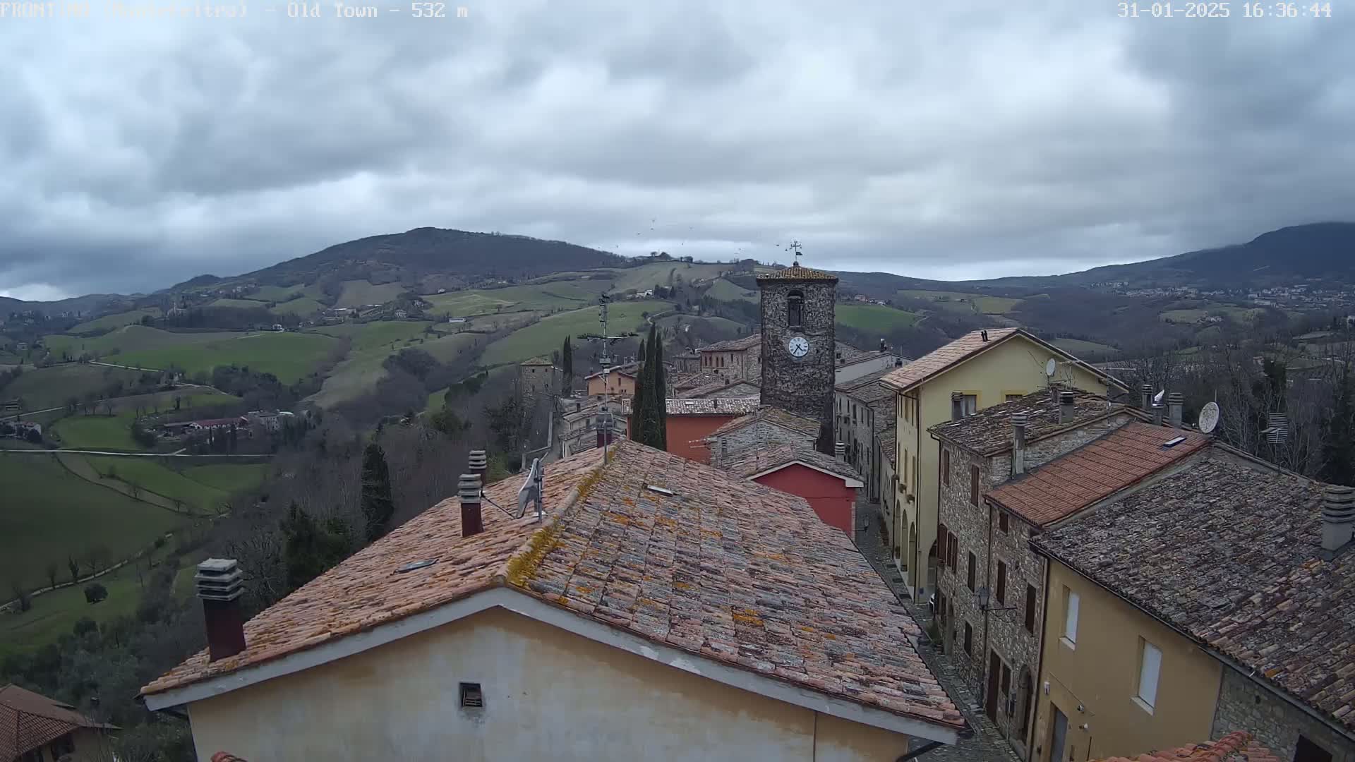 A stone village nestled in a valley overlooks rolling green hills under a cloudy sky.