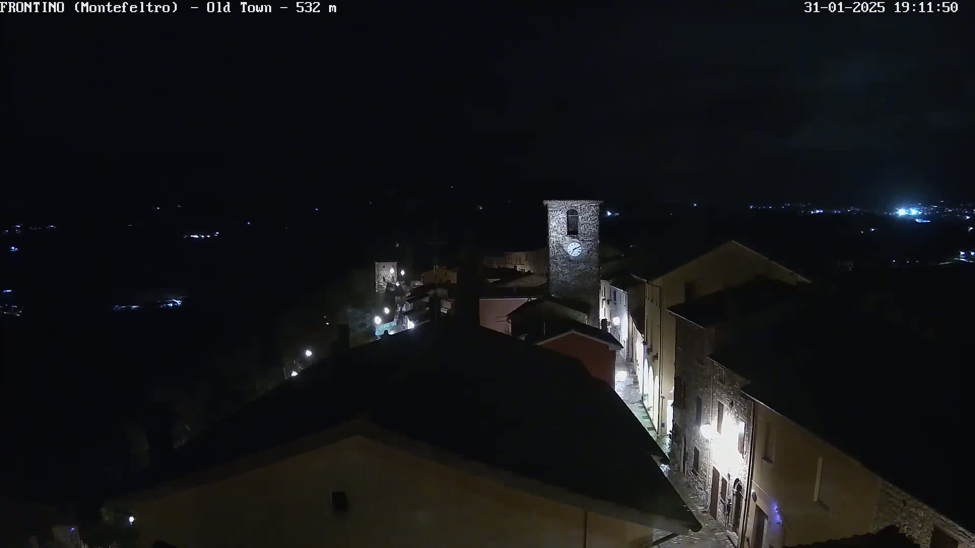 A nighttime, high-angle view shows a dimly lit stone village with a clock tower, under a dark, clear sky.