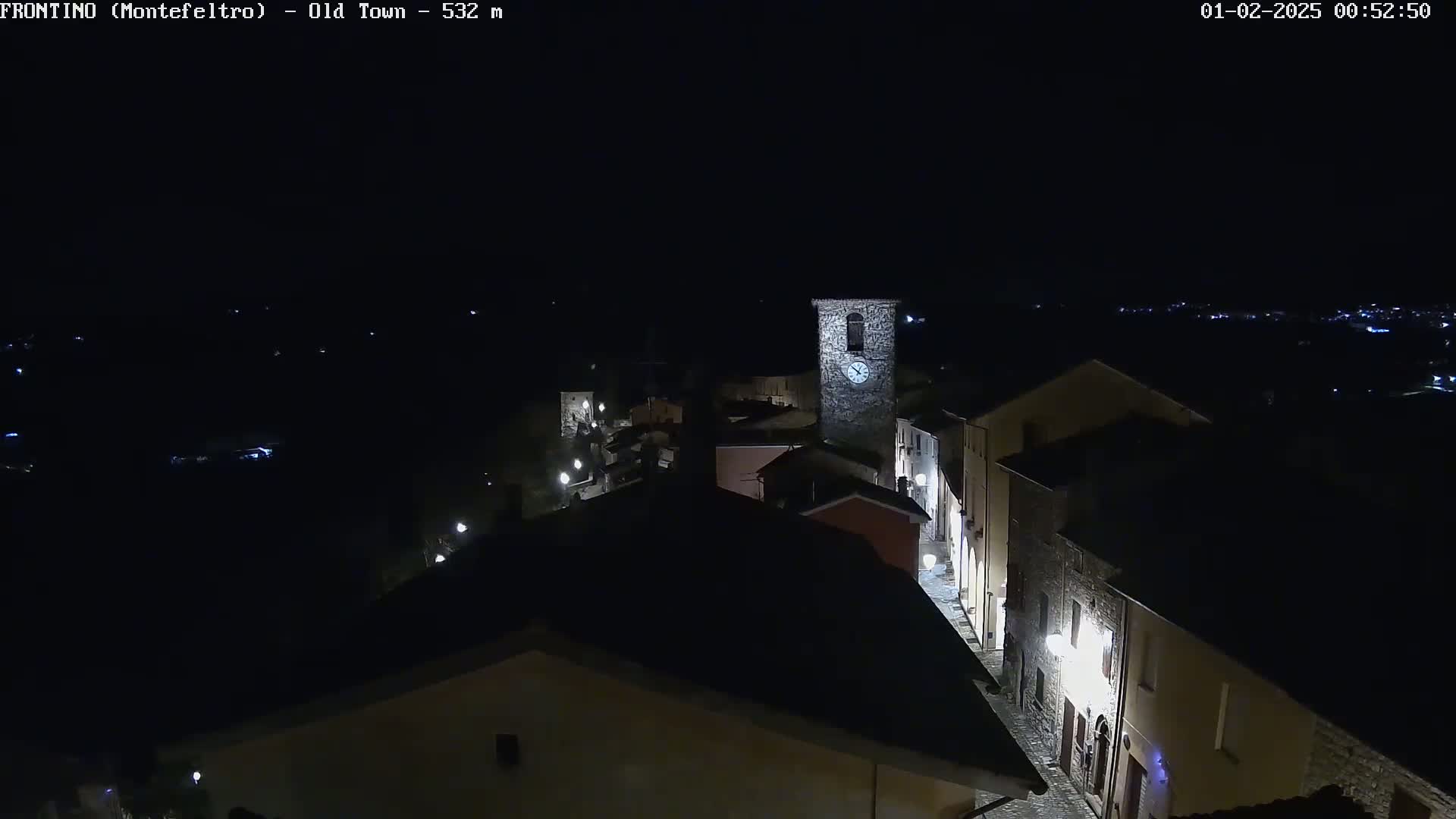 A nighttime aerial view shows a small, illuminated town with a clock tower, under a clear, dark sky.