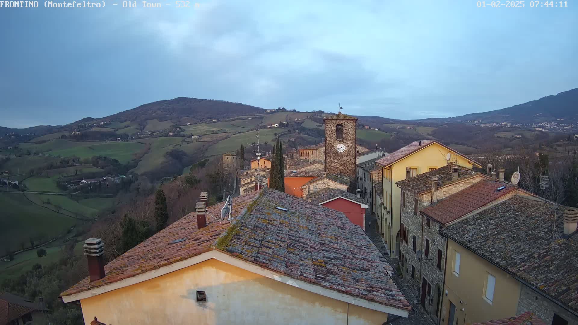 A view of a stone village nestled in rolling hills under a partly cloudy, dusky sky.
