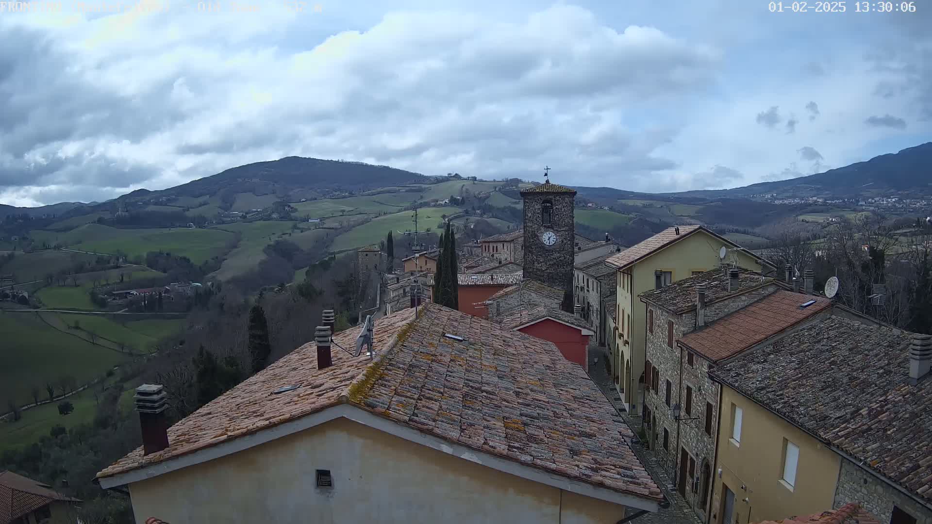 A hillside village of stone buildings with terracotta roofs is nestled in a valley under a partly cloudy sky.
