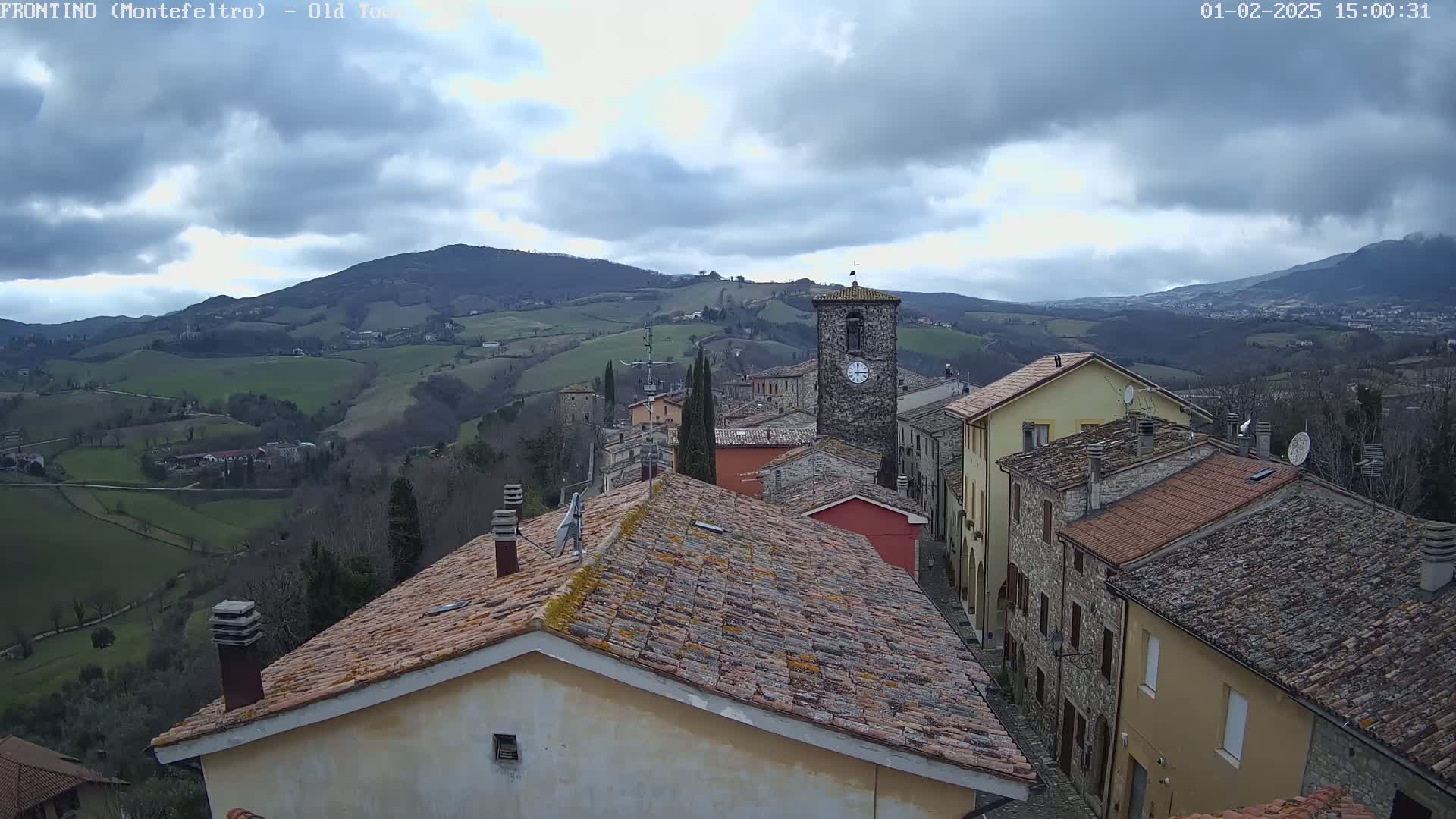 A clock tower stands prominently in a small village nestled in a valley, overlooked by rolling hills under a cloudy sky.
