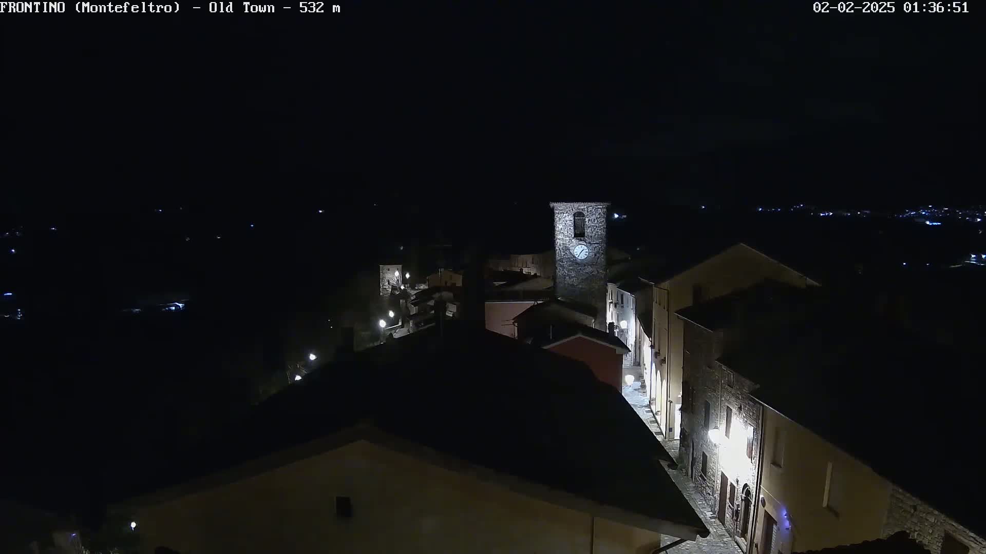 A nighttime aerial view shows a small, dimly lit town with a clock tower, under a clear, dark sky.