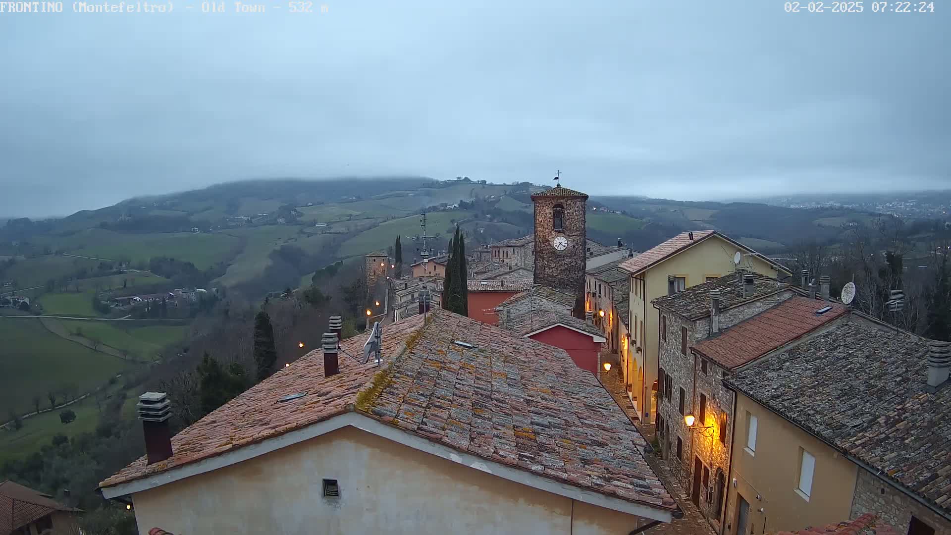 A hilltop village with terracotta roofs and a clock tower is nestled in a valley under an overcast sky.