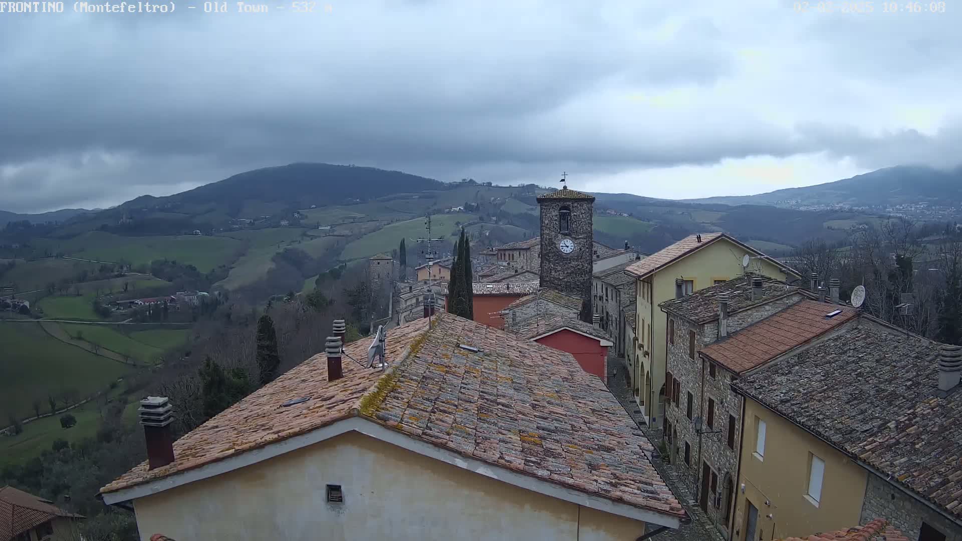 A stone village with a clock tower sits on a hillside overlooking a valley of rolling green fields under a cloudy sky.
