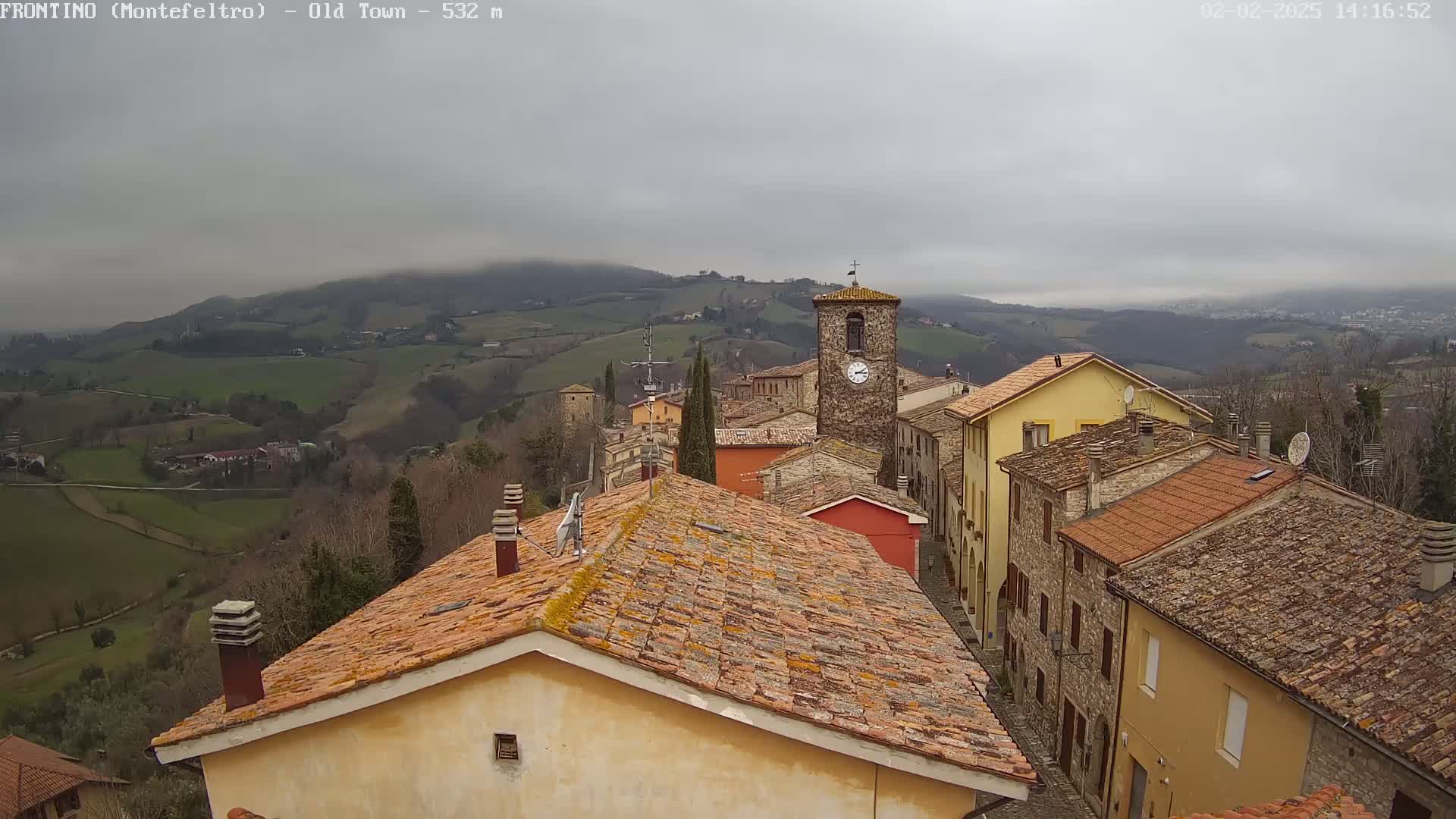 A clock tower and terracotta-roofed buildings in a hillside town are visible under an overcast sky.