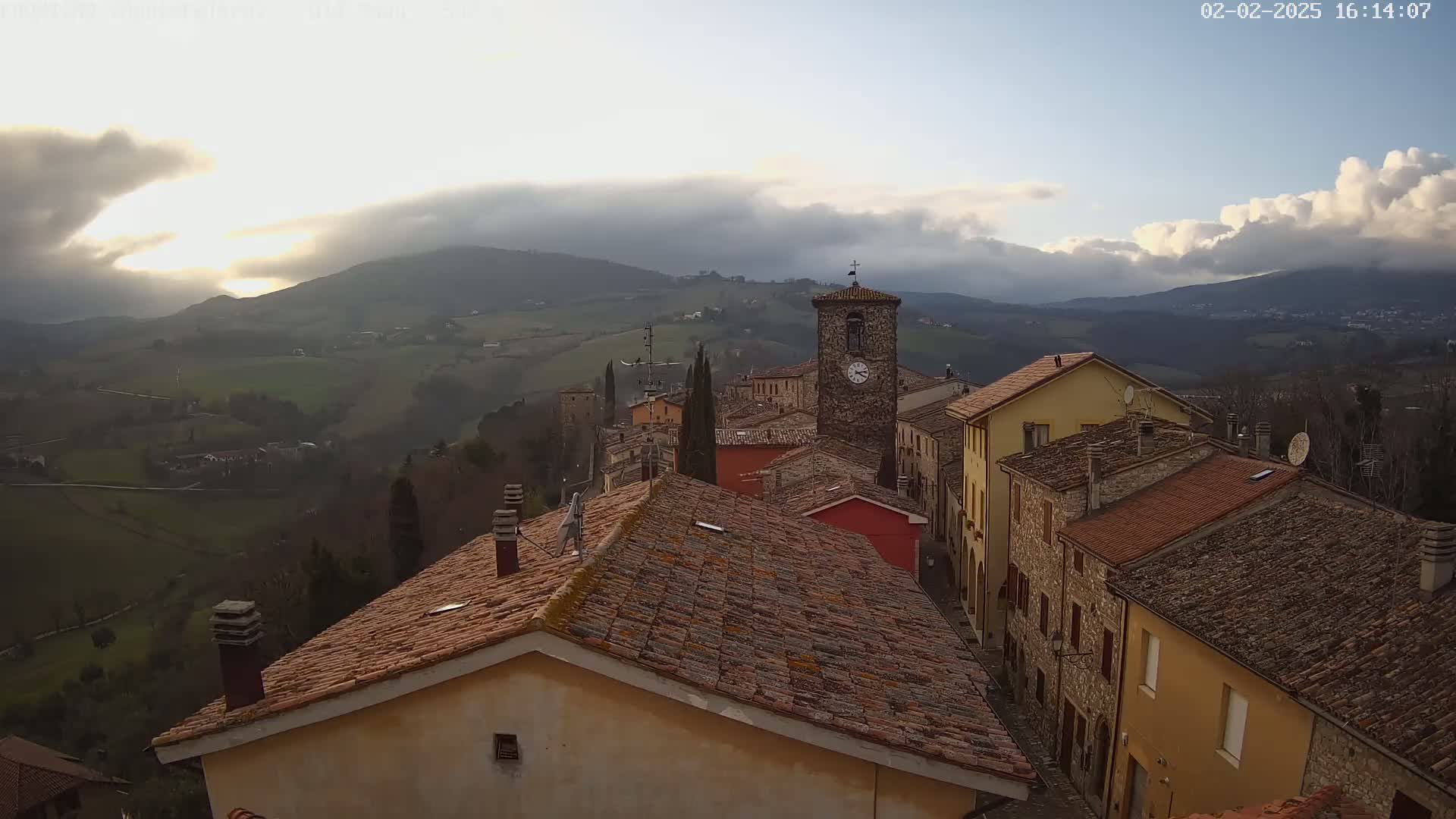 A partly cloudy day reveals a picturesque village nestled in rolling hills, with terracotta rooftops dominating the view.