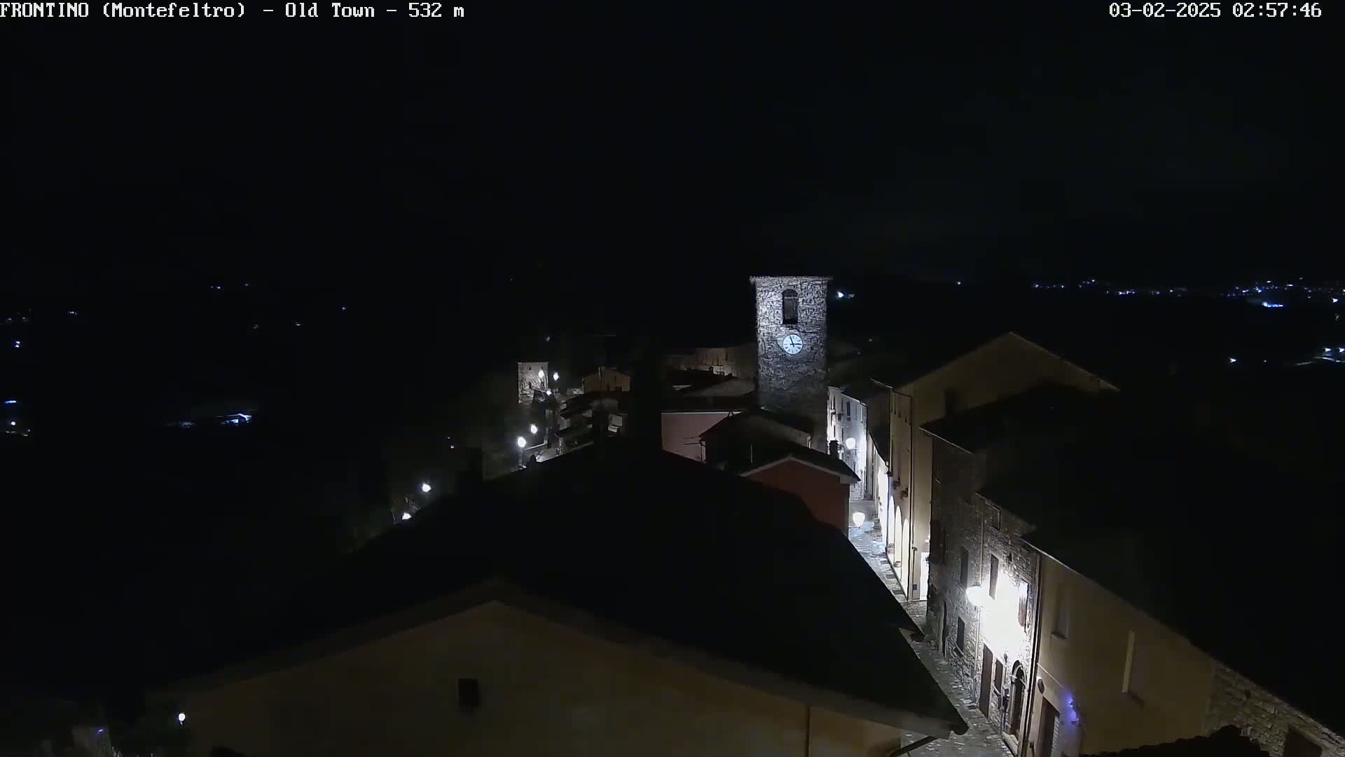 A nighttime, high-angle view shows a small, dimly lit town with a prominent stone clock tower, under a dark, clear sky.