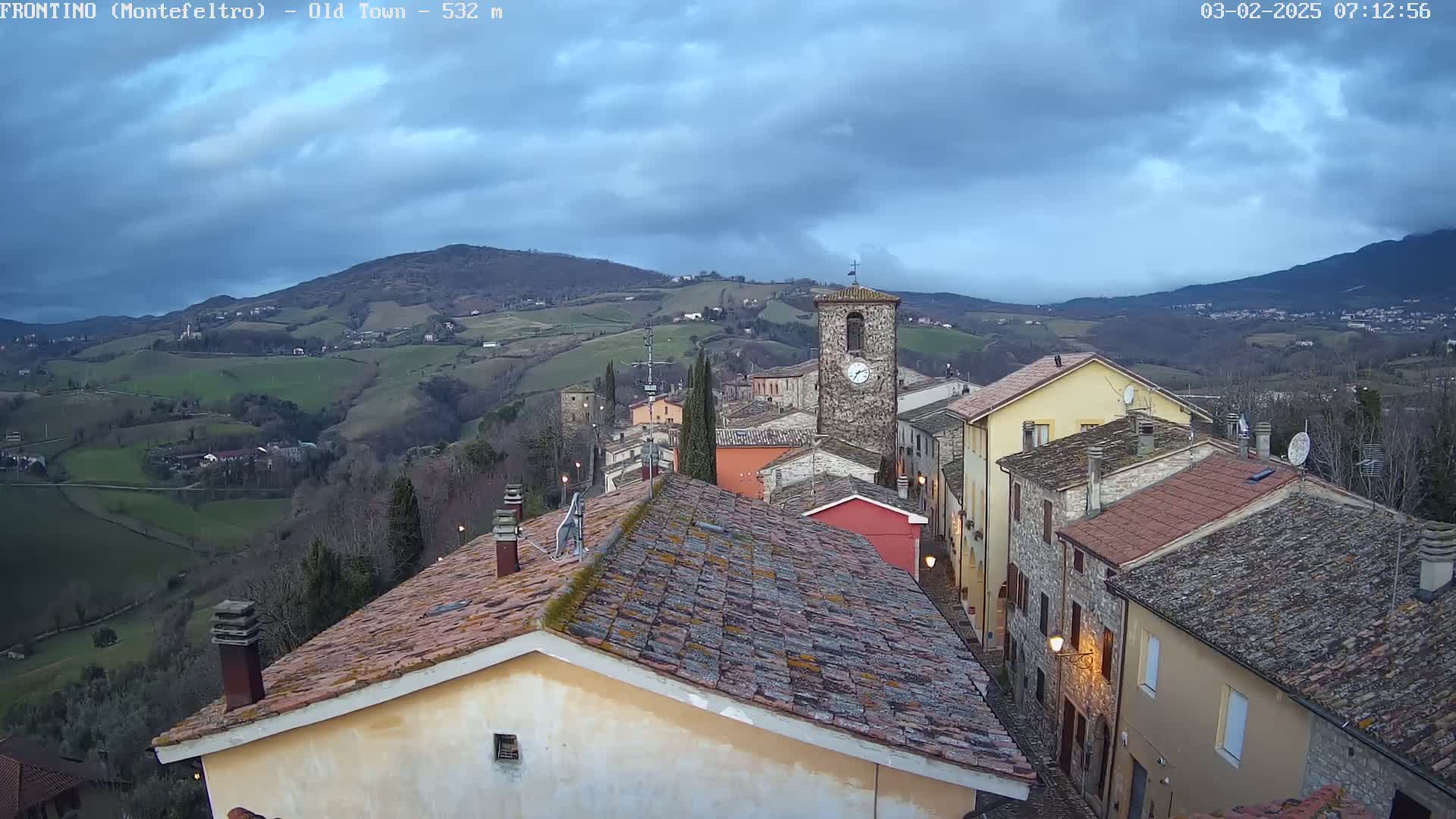 A small, hilly town with red-tiled roofs and a clock tower is visible under a cloudy sky.