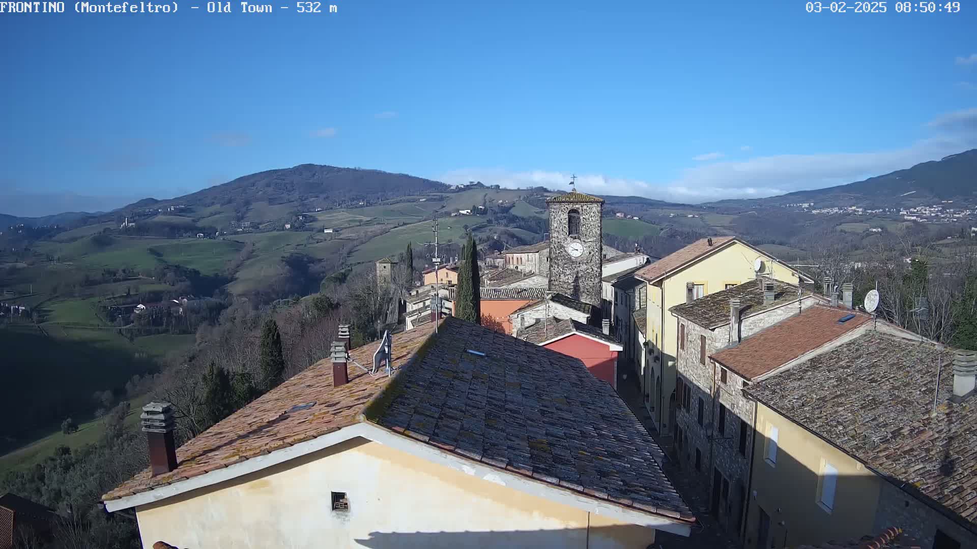 A picturesque Italian village nestled in rolling hills under a clear blue sky, with a clock tower as a prominent feature.