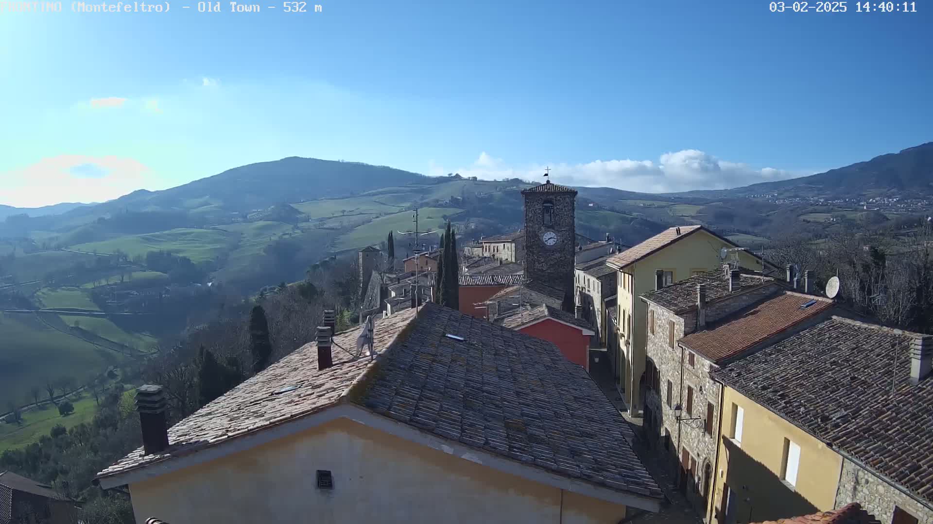 A sun-drenched village nestled in rolling green hills under a mostly clear blue sky.