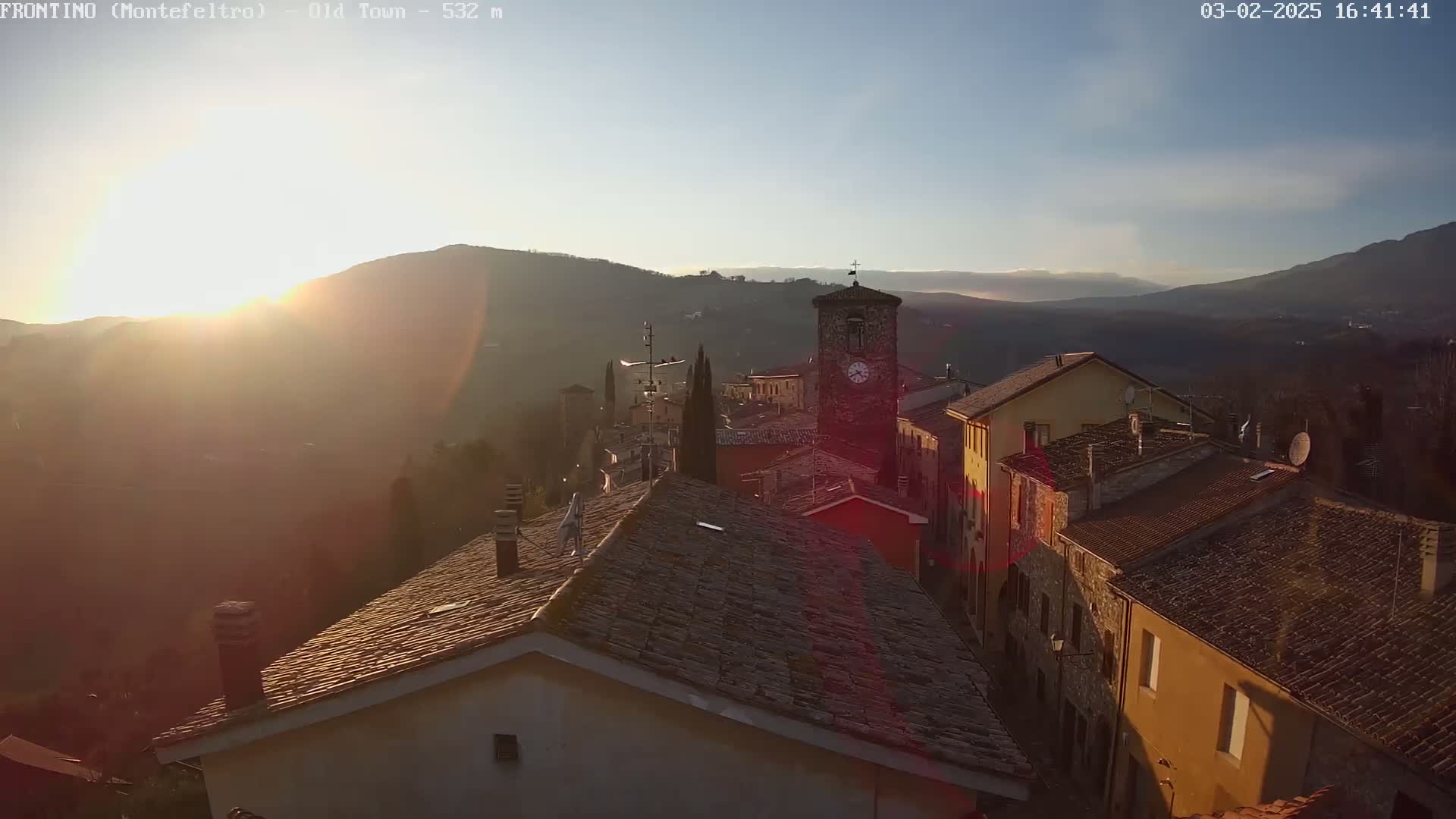 A sunlit village nestled in a valley between hills, with a clock tower prominent in the scene.