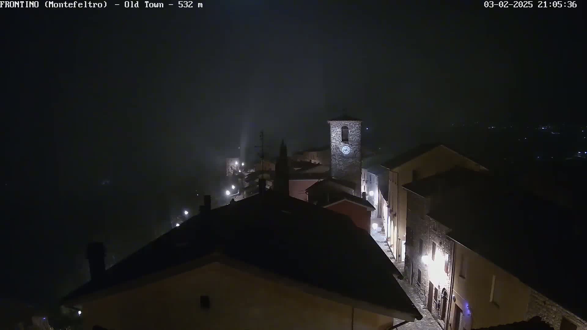 A nighttime view of a fog-shrouded town with stone buildings and streetlights, a clock tower visible in the center.