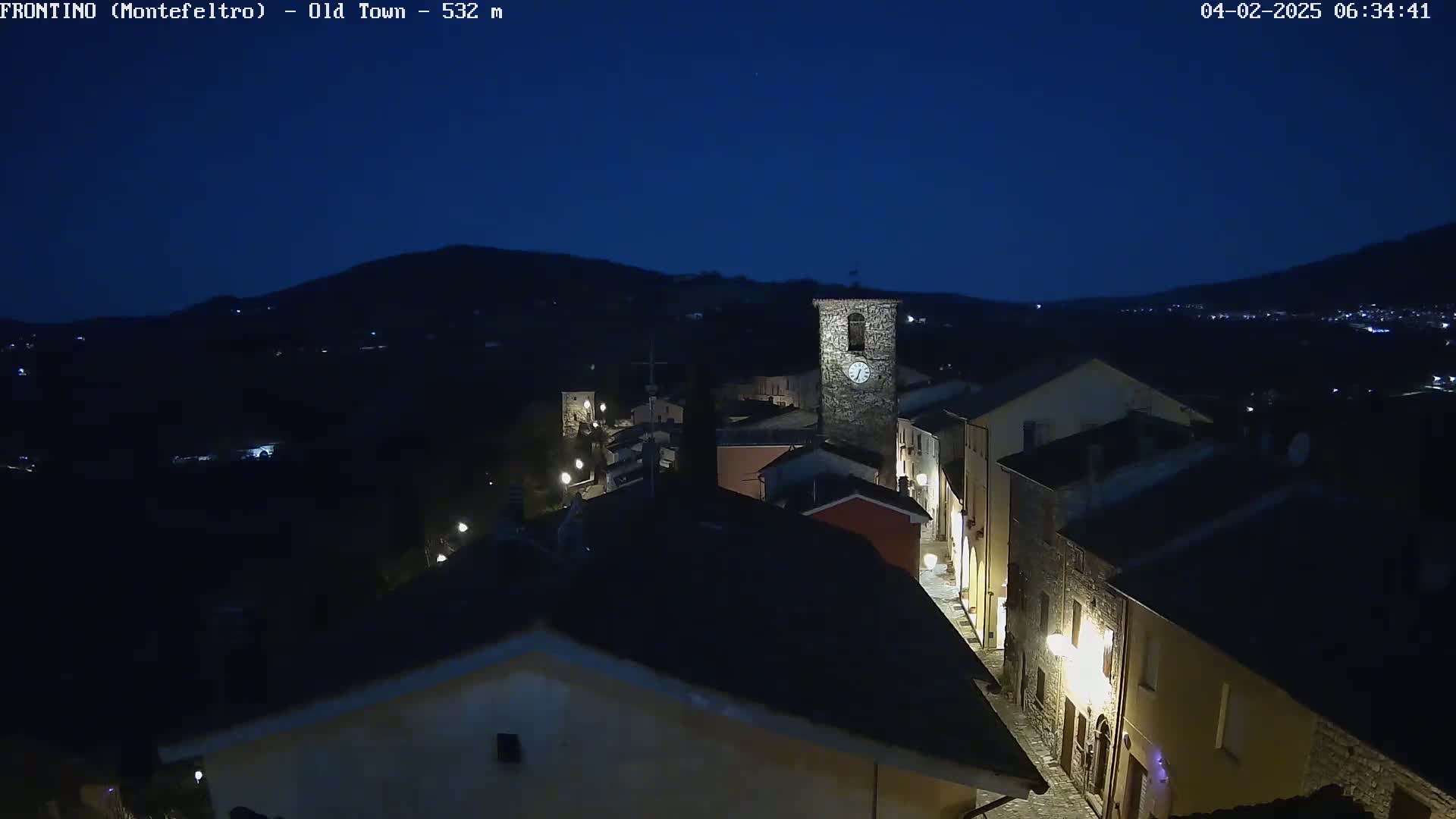 A nighttime, high-angle view shows a dimly lit stone village nestled in a valley under a dark blue sky.