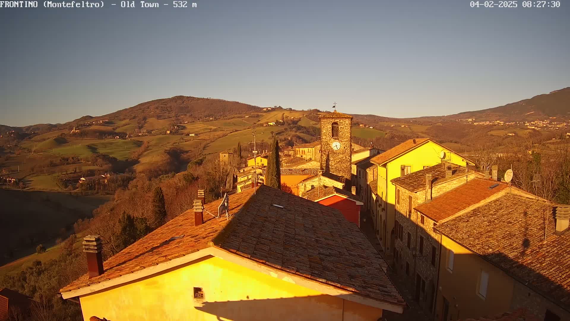 A sunlit hillside village with terracotta roofs and a clock tower, nestled amongst rolling hills under a clear, pale blue sky.