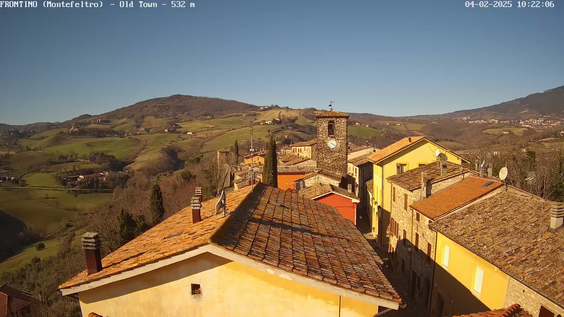 A sunny day reveals a quaint village nestled in a rolling hillside, with terracotta rooftops and a prominent clock tower.