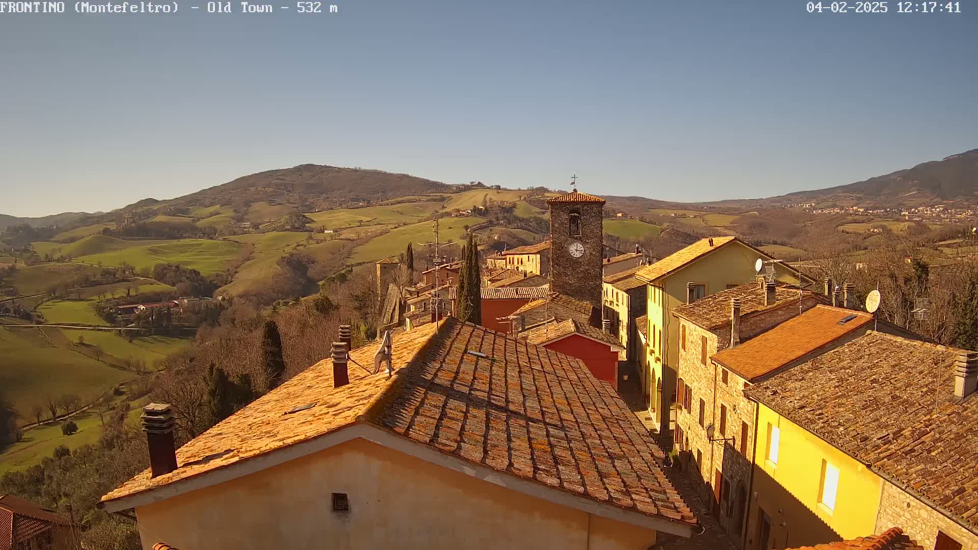 A sunlit village of stone buildings with terracotta roofs nestles in a valley, overlooked by rolling green hills under a clear blue sky.