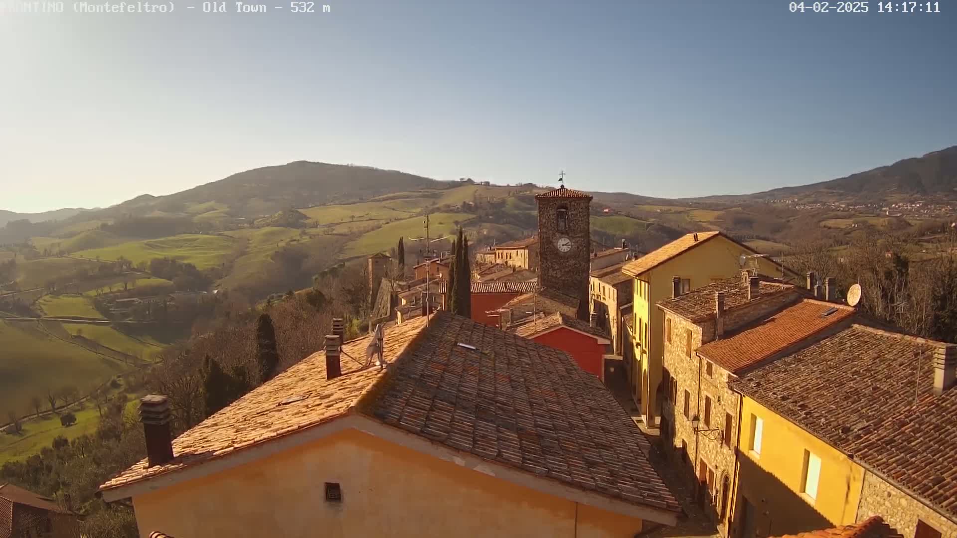 A sunlit Italian village with terracotta roofs nestles in a valley, surrounded by rolling green hills under a clear blue sky.