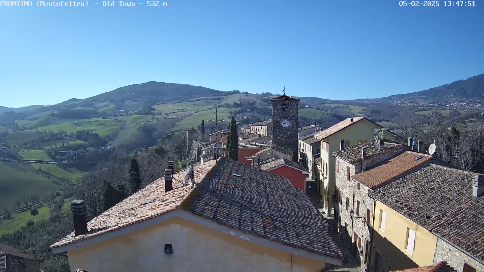 A clock tower stands in the center of a small village nestled in a valley, under a clear blue sky on a sunny day.
