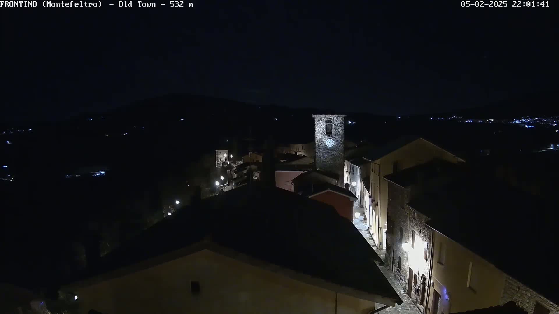 A nighttime aerial view shows a dimly lit stone village street with a clock tower, under a dark, clear sky.
