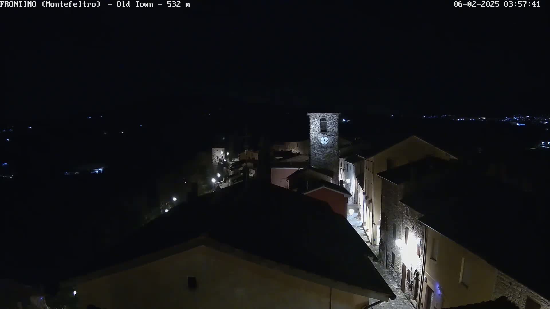 A nighttime aerial view shows a small town with stone buildings, lit streets, and a clock tower, under a clear, dark sky.