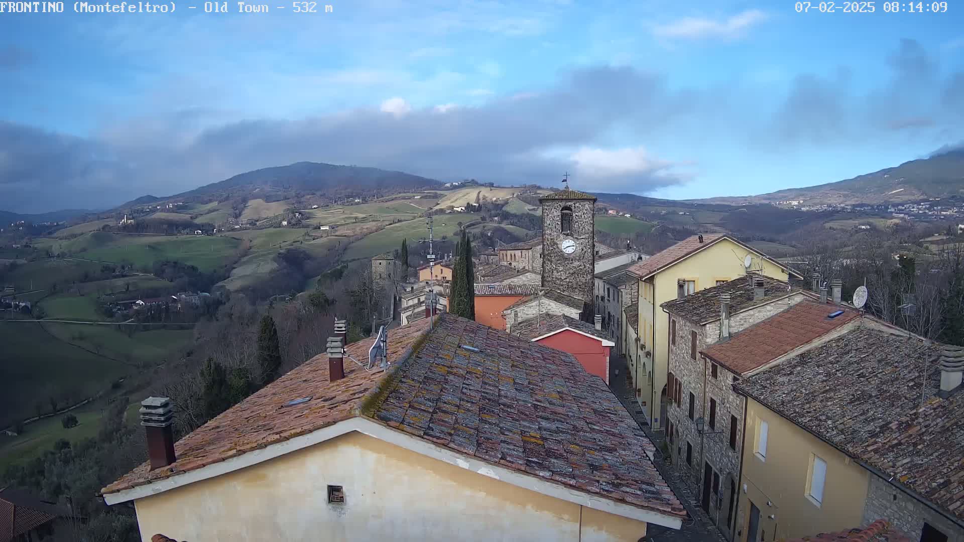 A small village nestled in a valley, under a partly cloudy sky, is viewed from above, showing rooftops and a clock tower.
