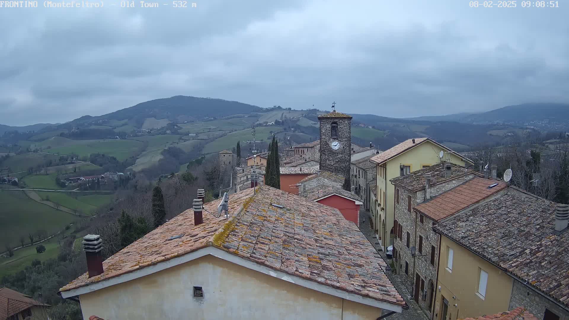 An overcast day reveals a quaint village nestled in a valley, its stone buildings and terracotta rooftops overlooking rolling green hills.