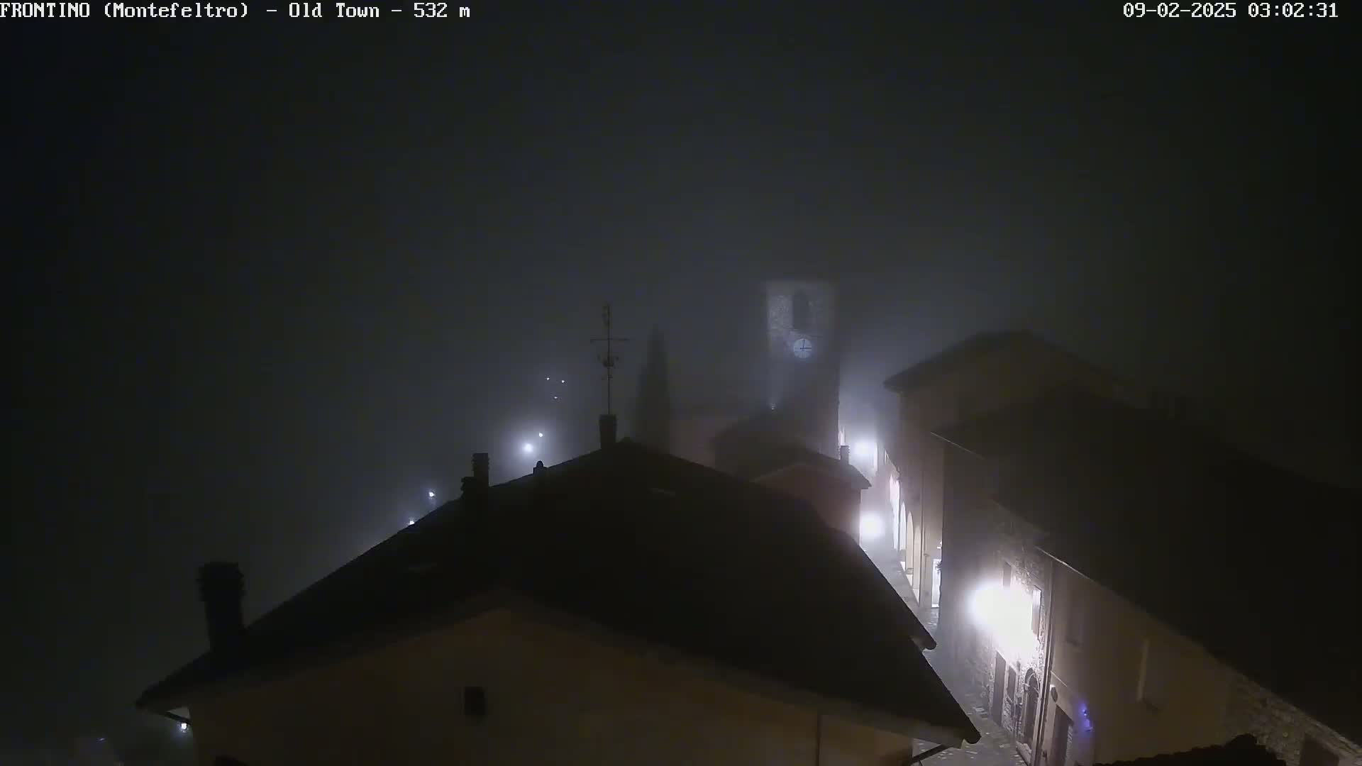 A foggy night scene shows the rooftops of a town, with a clock tower visible in the distance under dimly lit streetlights.