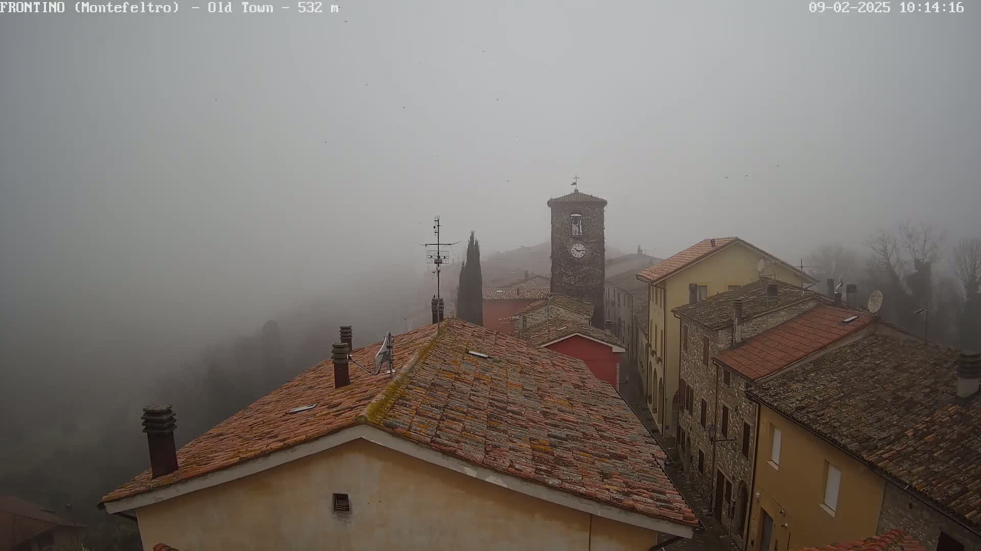 A foggy view of a town's rooftops and a clock tower, showing stone and tile buildings.